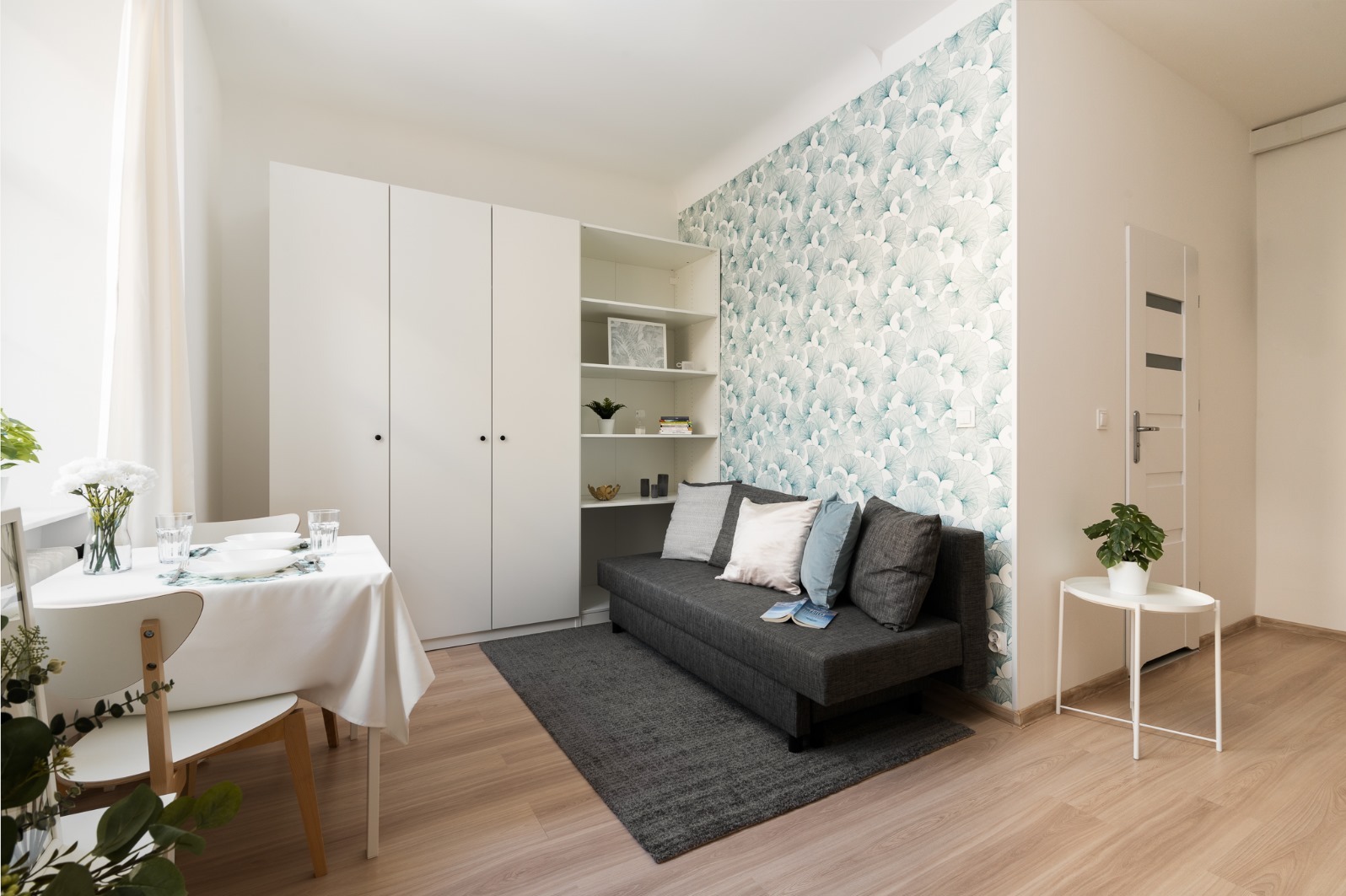 Jasne wnętrze pokoju z sofą na tle ściany w delikatny, niebiesko-biały wzór, obok biała szafa, stół z nakryciem i kwiatami, oraz mały stolik z rośliną doniczkową.