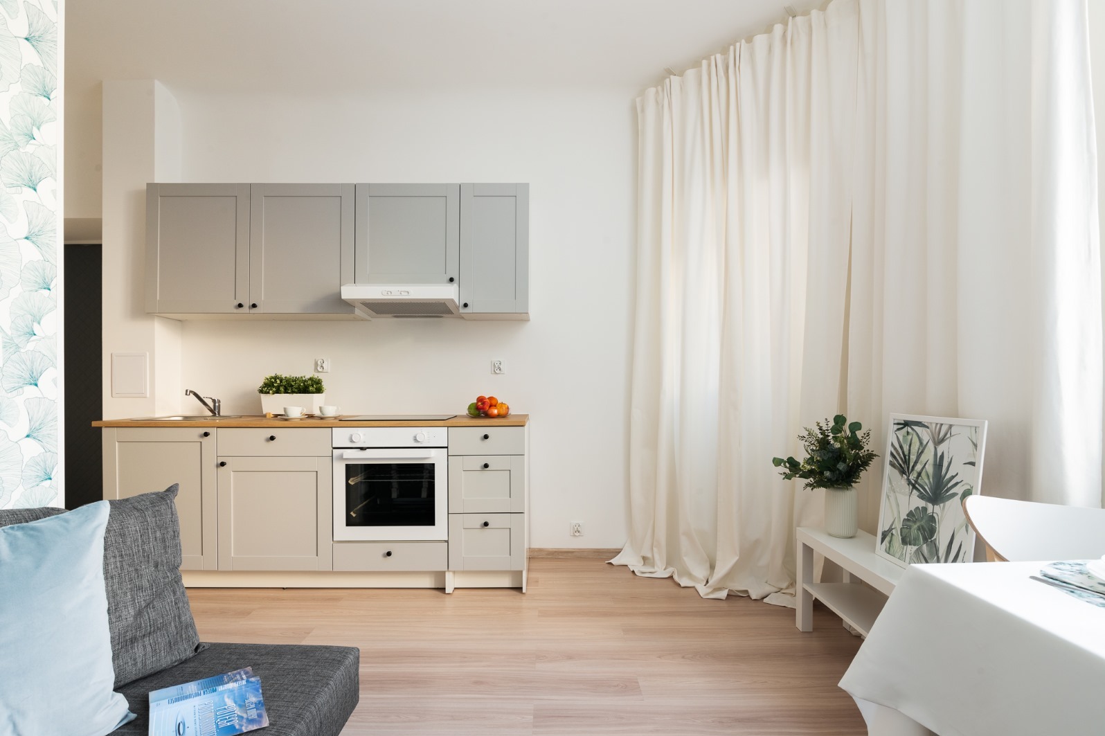 Jasna kuchnia z szarymi szafkami i drewnianym blatem, widok z salonu z szarą sofą i dekoracyjną poduszką, białe zasłony w tle.