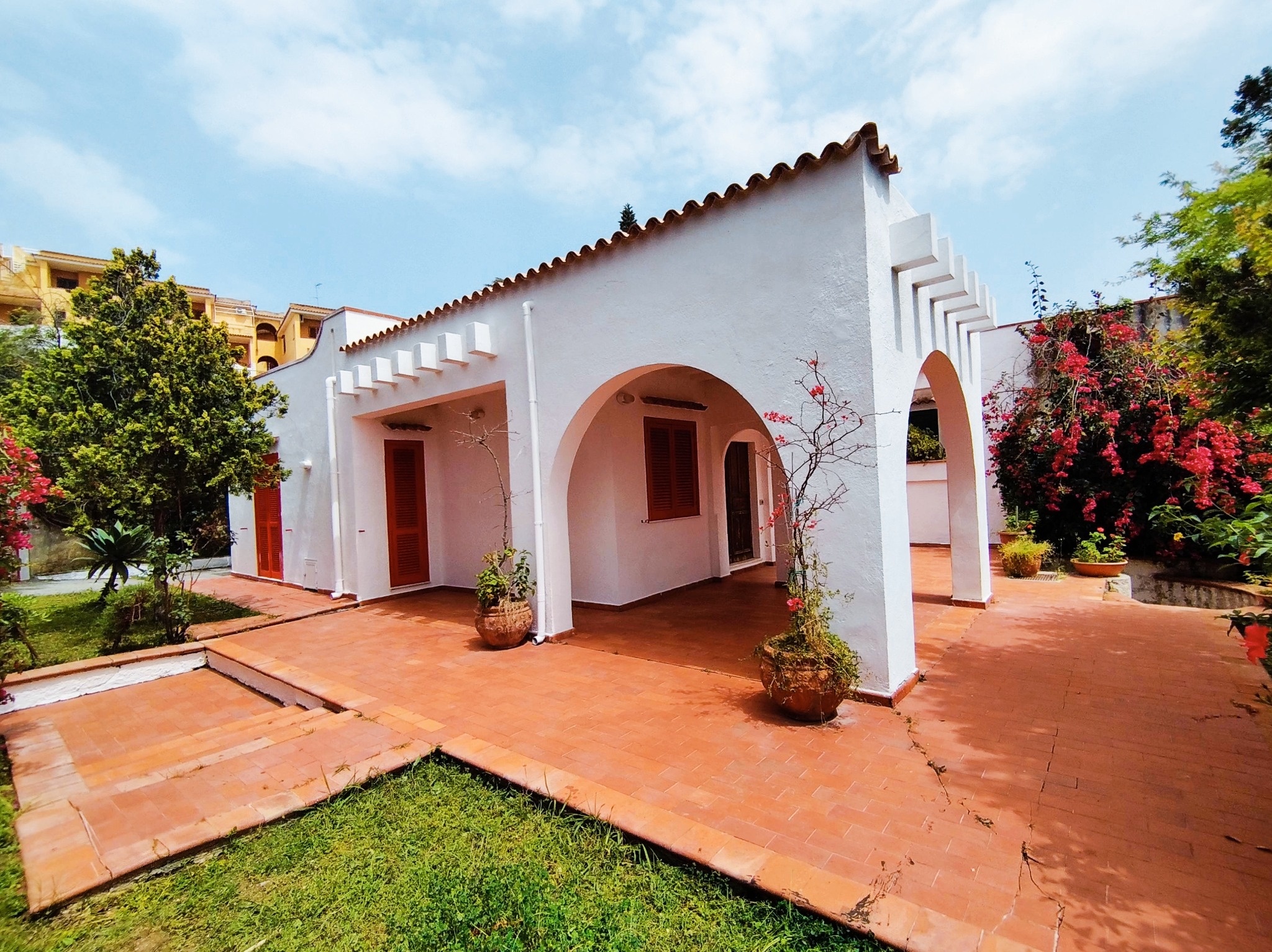 Biały budynek z łukami i terakotową podłogą, rośliny w donicach, widok na ogród i błękitne niebo z lekkimi chmurami.