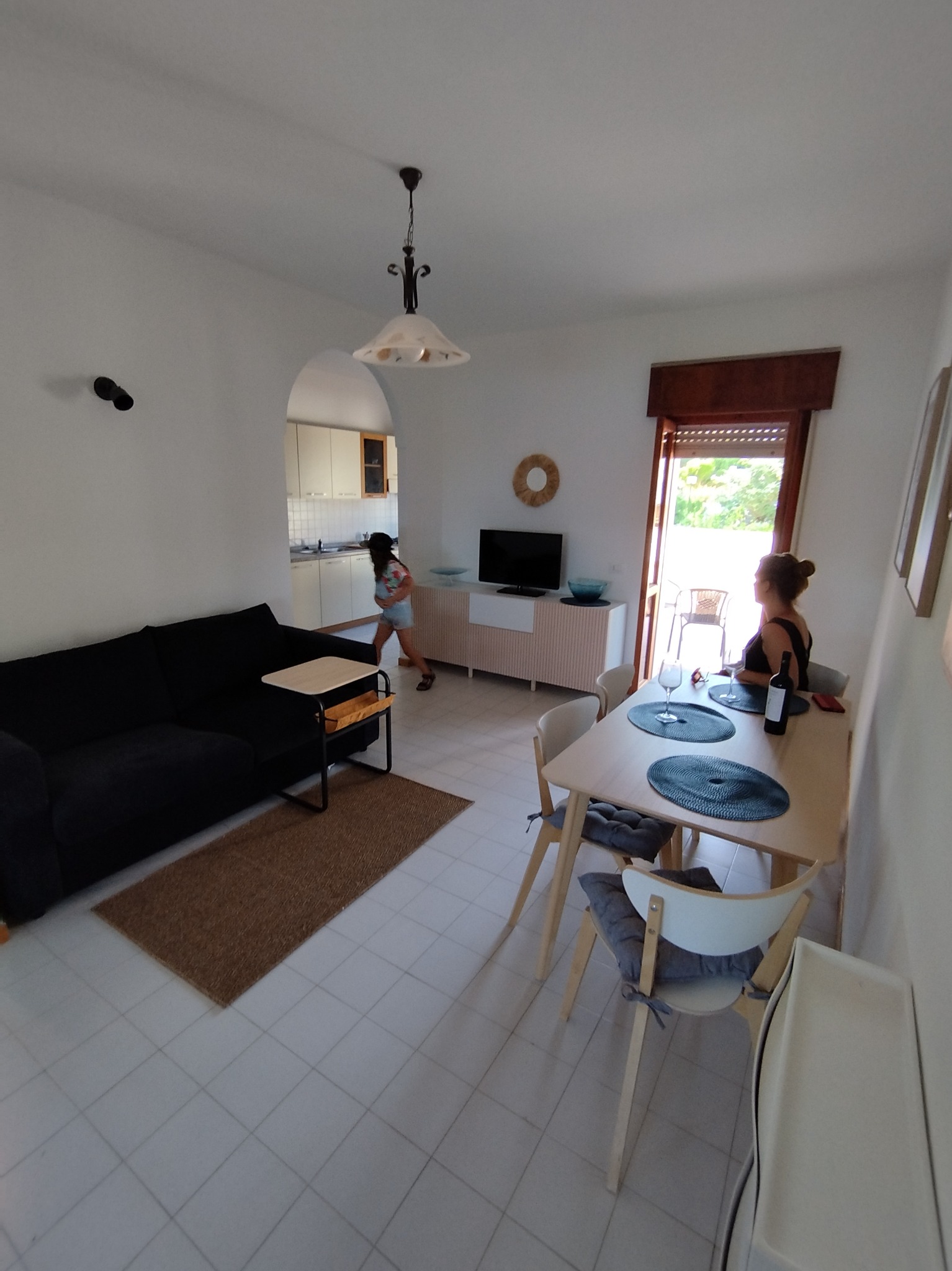 Jasne wnętrze mieszkania z czarną sofą, drewnianym stołem jadalnym z winem, telewizorem i widokiem na taras z ogrodem przez otwarte drzwi balkonowe