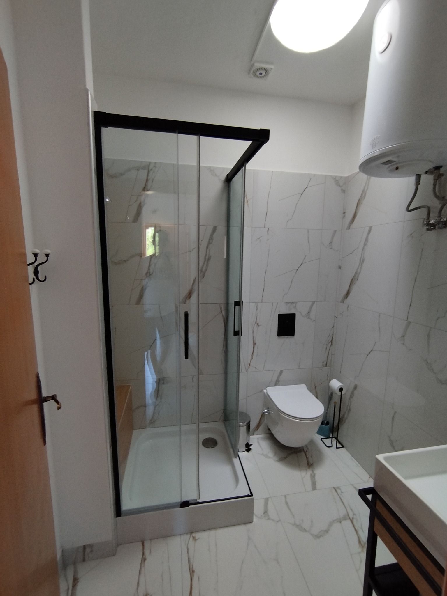 Nowoczesna łazienka z kabiną prysznicową typu walk-in z czarnymi ramami, marmurowe płytki na ścianach i podłodze, podwieszana toaleta i biały bojler.