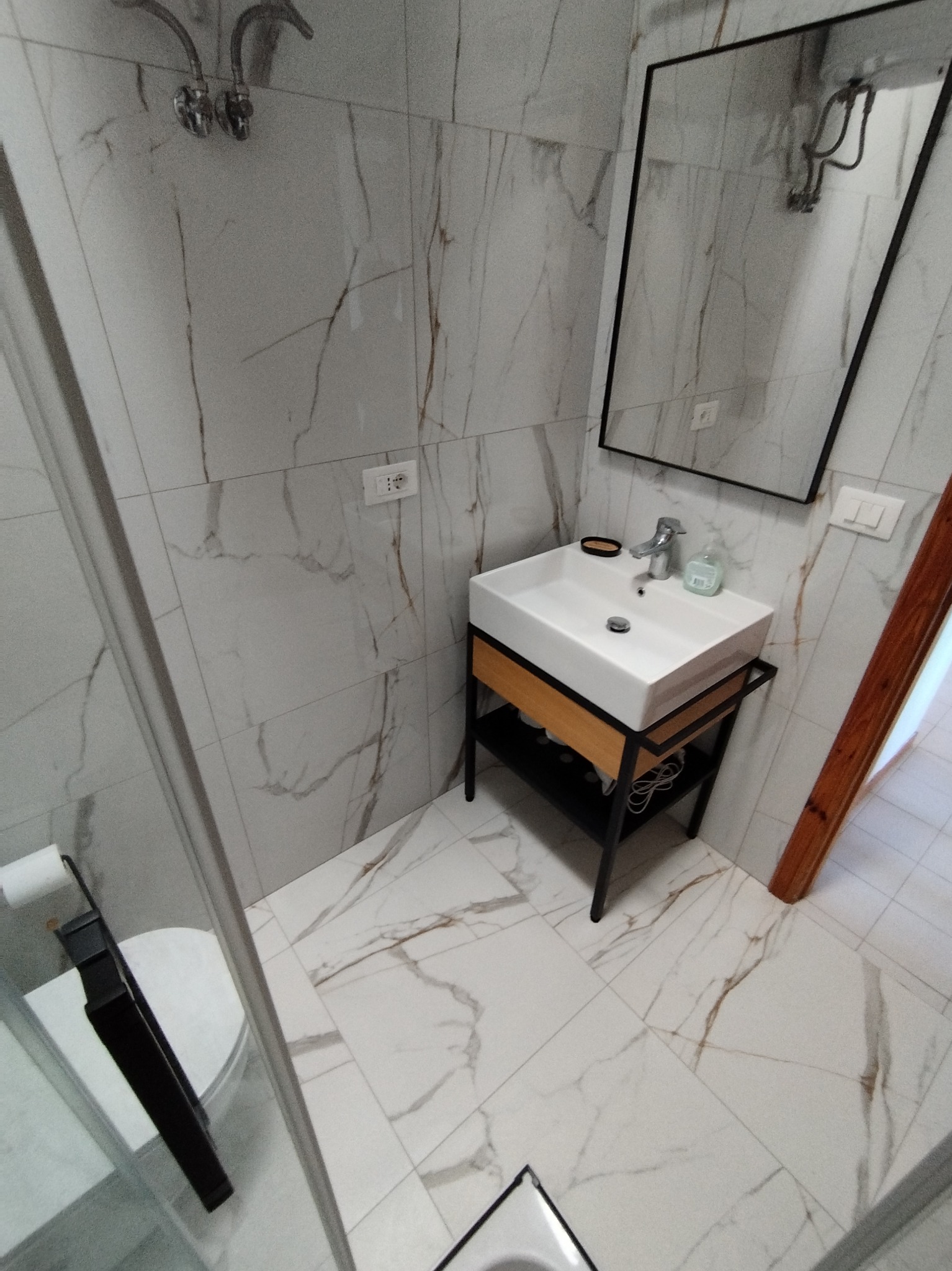 Nowoczesna łazienka z białą umywalką na metalowym stelażu z drewnianymi elementami, lustrem w czarnej ramie i płytkami imitującymi marmur na ścianach i podłodze.