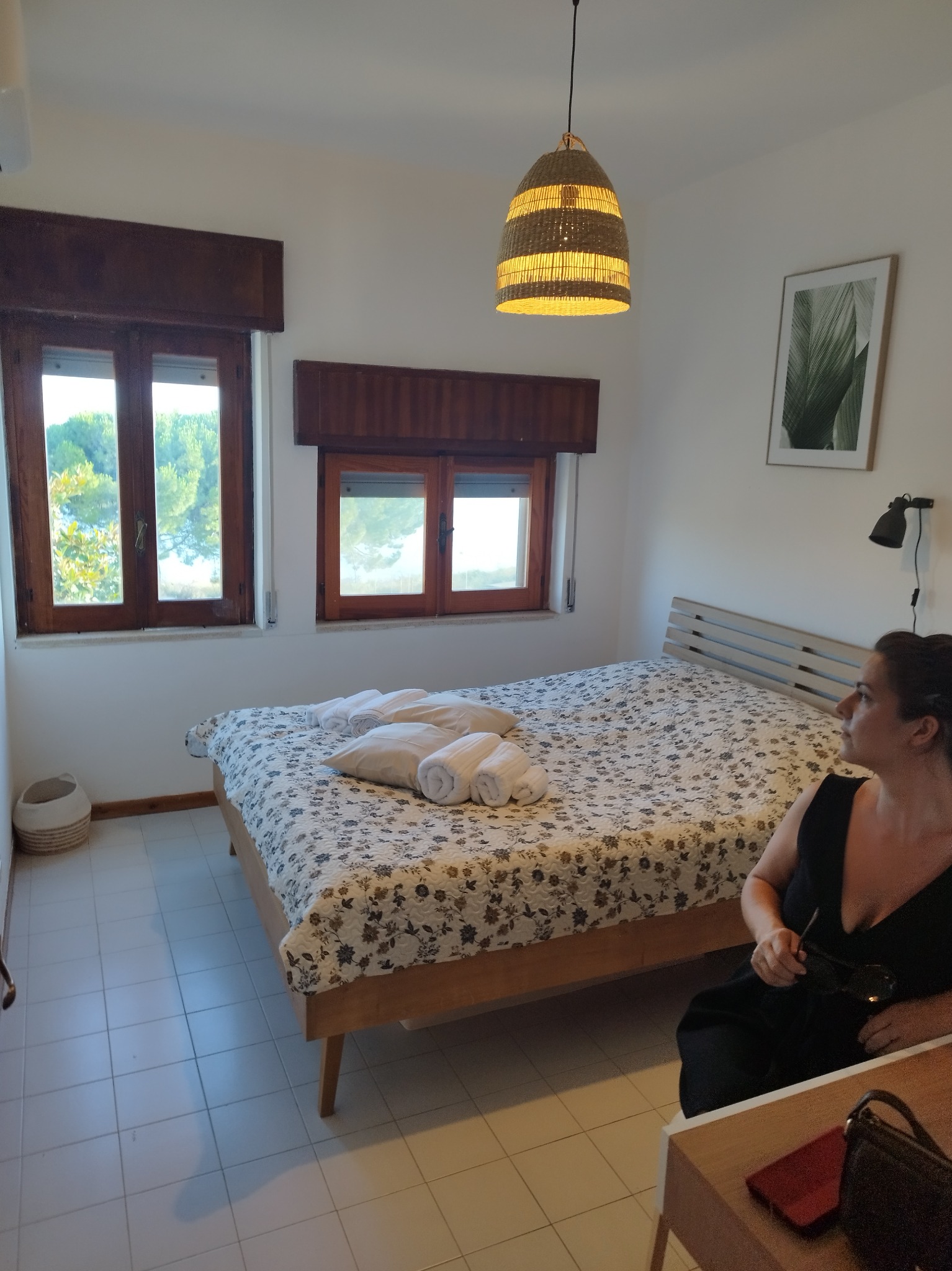 Jasna sypialnia z łóżkiem w kwiaty, drewnianymi oknami z widokiem na zieleń i osobą siedzącą na krześle. Wnętrze dopełnia wiklinowa lampa i minimalistyczny obraz z motywem roślinnym.