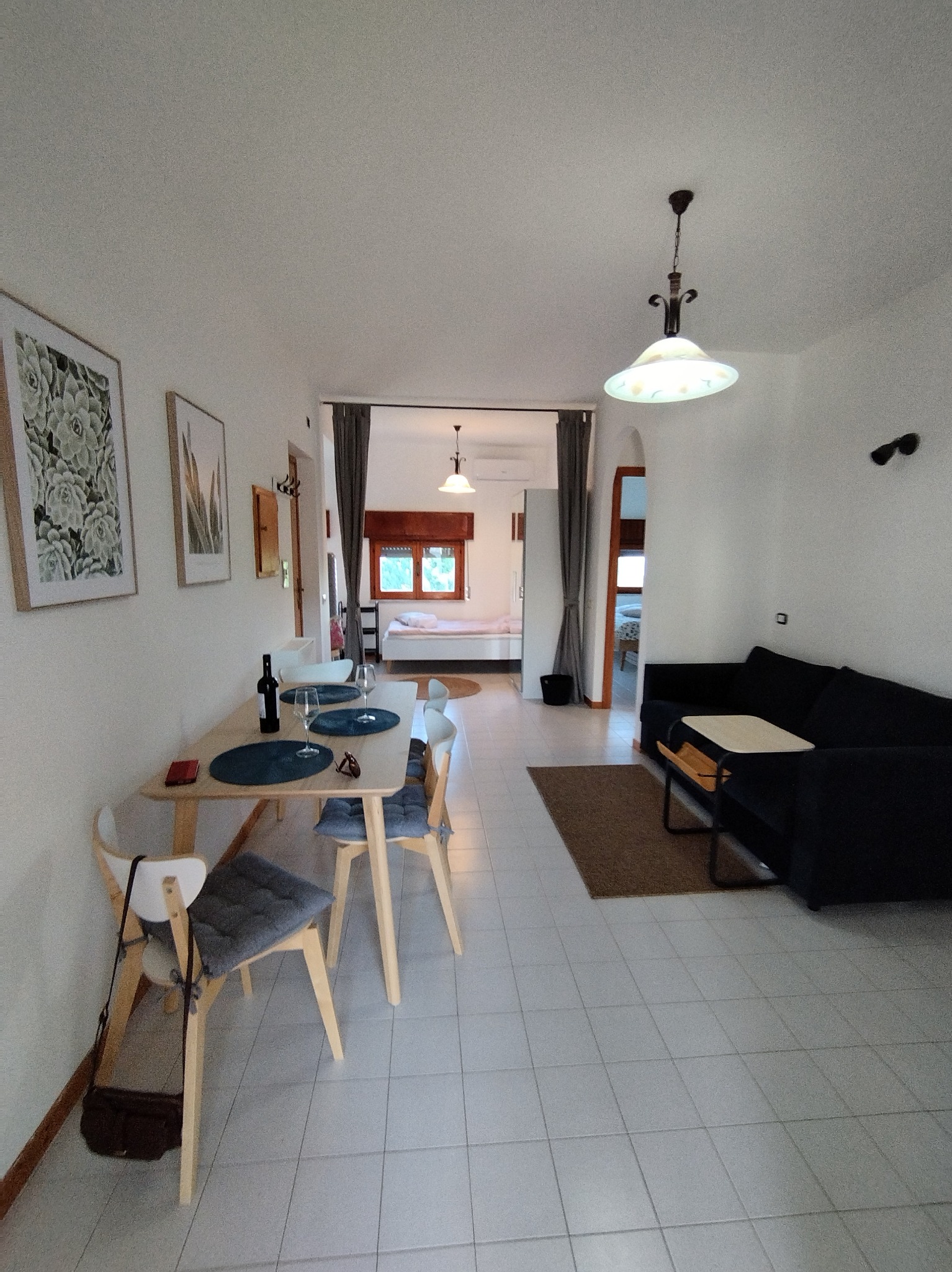 Jasne, przestronne wnętrze mieszkania z widokiem na sypialnię, stół z nakryciem dla dwojga, czarna sofa i lampa wisząca w stylu retro.