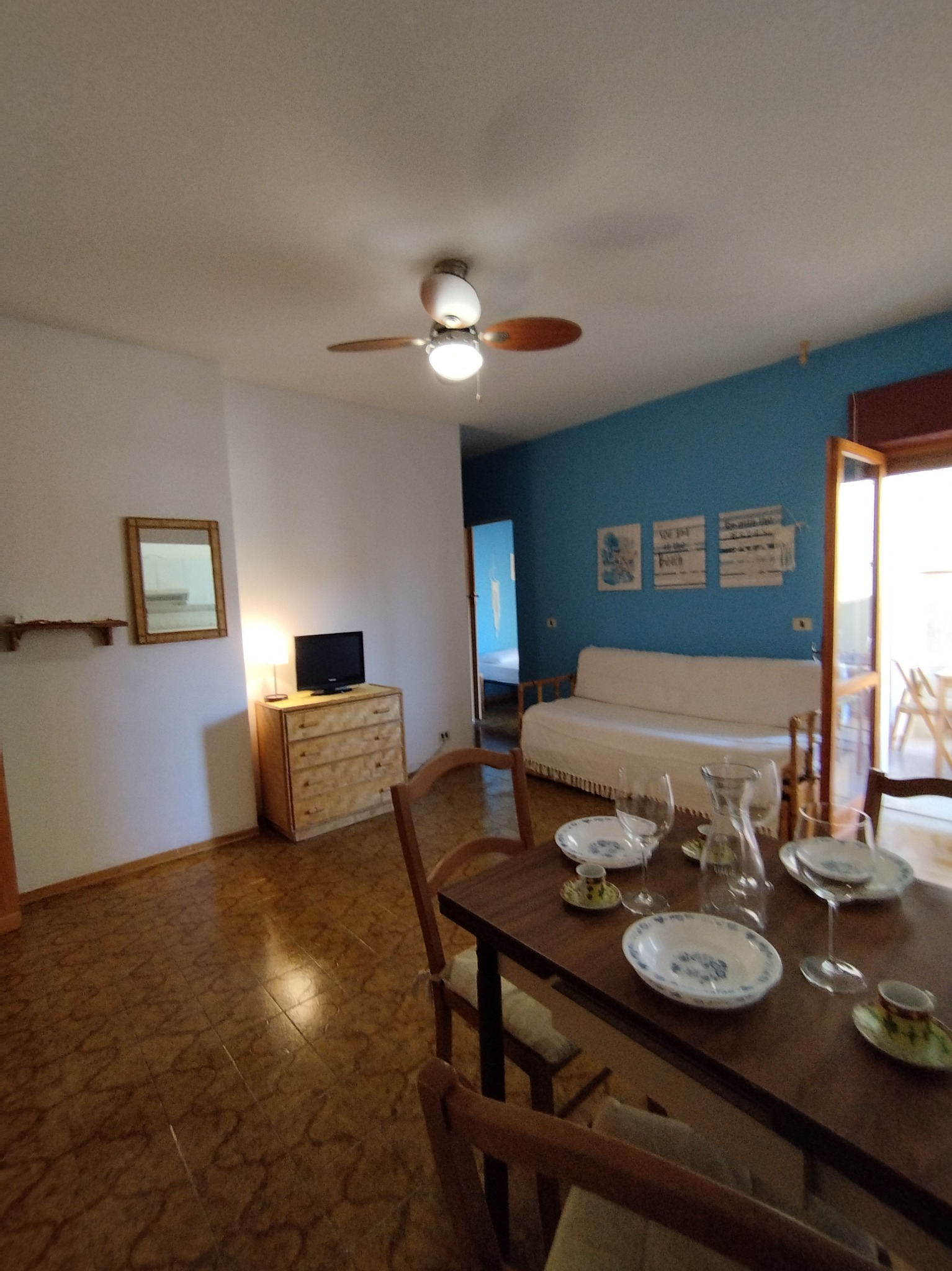 Jasne wnętrze jadalni z drewnianym stołem nakrytym porcelanową zastawą w niebieskie wzory, widok na salon z turkusową ścianą i kanapą, oraz otwarte drzwi balkonowe z widokiem na zewnątrz.