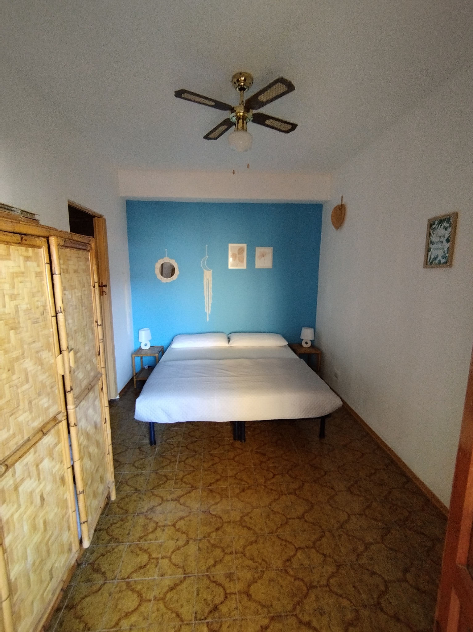 Pokój z łóżkiem na metalowej ramie, niebieską ścianą z dekoracjami, bambusowym parawanem i wzorzystą podłogą, oświetlony lampą z wentylatorem sufitowym.