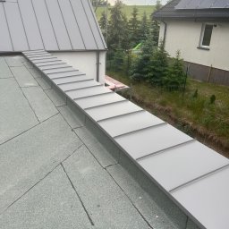 M-bart Dach - Szara obróbka blacharska na styku dachu pokrytego papą i ściany budynku, widok z góry na tle zieleni i sąsiedniego domu z panelami słonecznymi.