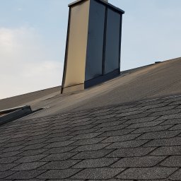 M-bart Dach - Nowy, błyszczący komin z obróbką blacharską na dachu pokrytym gontem bitumicznym w odcieniach szarości, widoczne łączenie z połacią dachu.