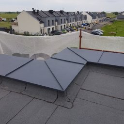 M-bart Dach - Szary dach z papy z obróbkami blacharskimi w kształcie rombów na tle szeregu domów jednorodzinnych i pochmurnego nieba.