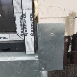 M-bart Dach - Szczegółowe ujęcie obróbki blacharskiej okna na dachu z widocznymi śrubami mocującymi i pianką uszczelniającą, fragment dachu pokrytego papą oraz ramą okna z folią ochronną producenta Krispol.