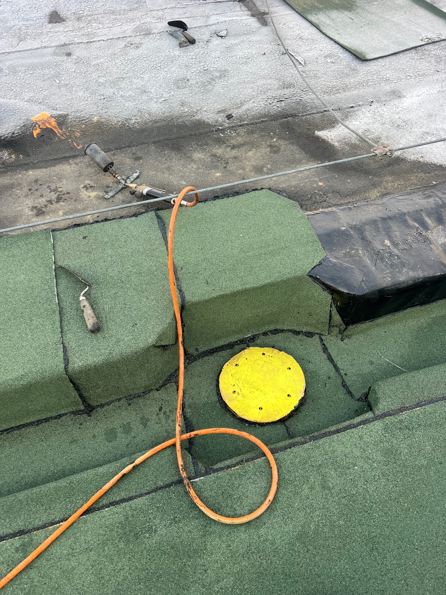 Prace dekarskie na płaskim dachu: zielona papa termozgrzewalna, narzędzia dekarskie (palnik z płomieniem, kielnia), żółty znacznik i pomarańczowy wąż gazowy.