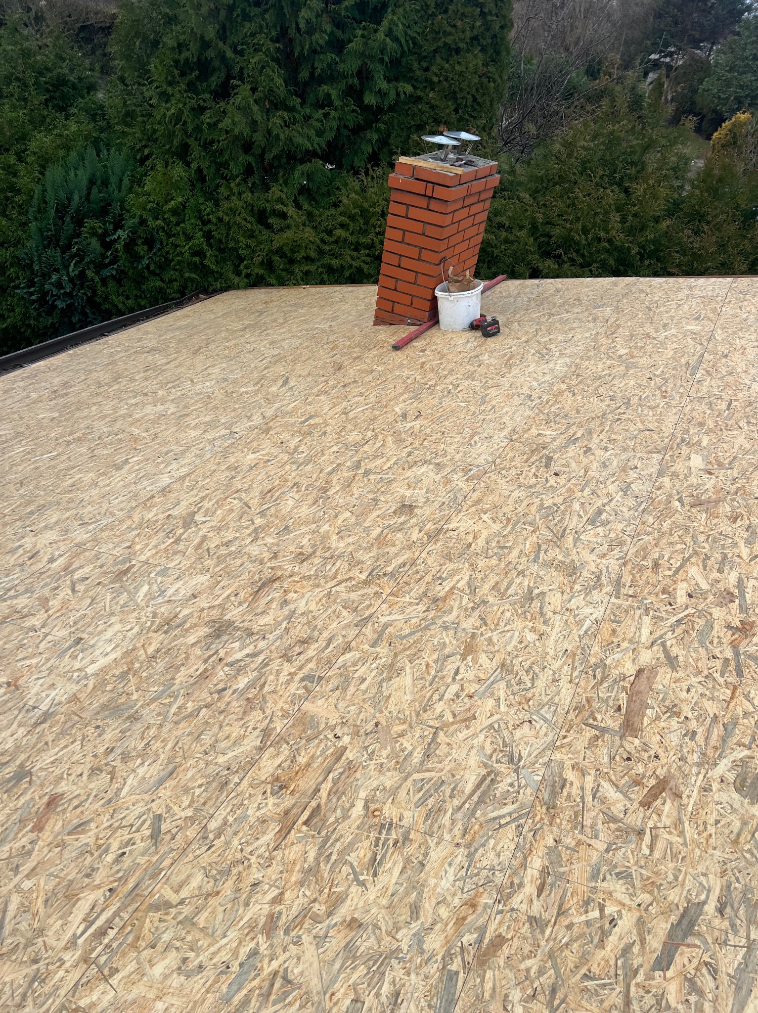Widok z góry na przygotowaną podbudowę dachu z płyt OSB, z kominem w tle, wiaderkiem i wkrętarką akumulatorową, otoczoną zielonymi drzewami.