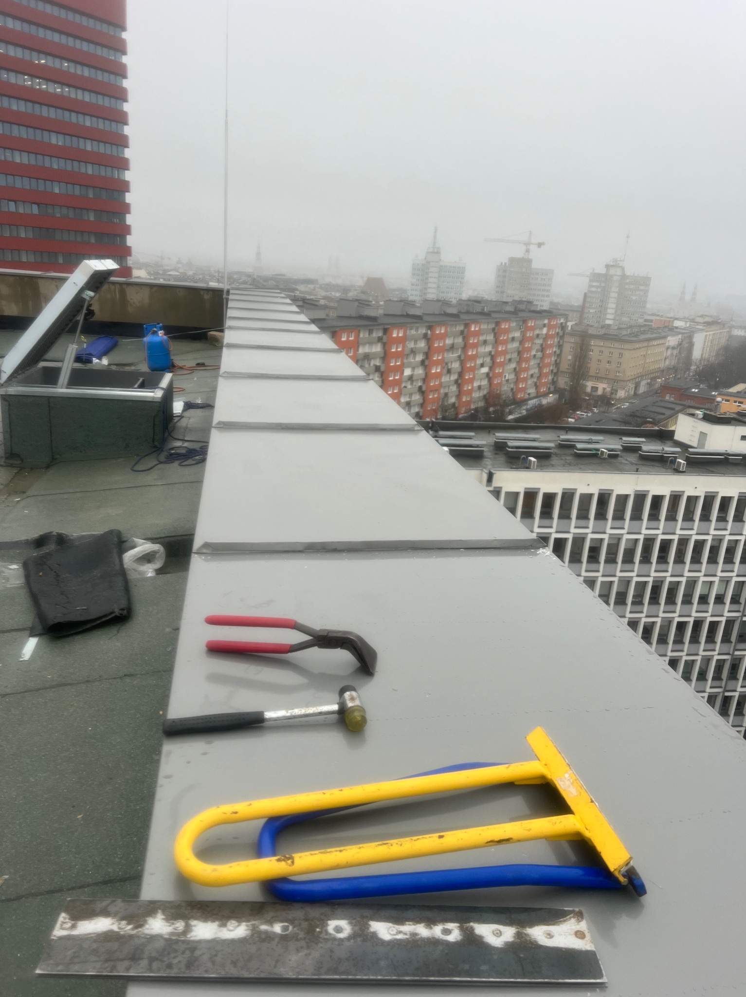 Blaszany dach z narzędziami dekarskimi (szczypce, młotek, giętarka) na tle miejskiego krajobrazu w mglisty dzień. Widoczny fragment instalacji solarnej i butla gazowa.