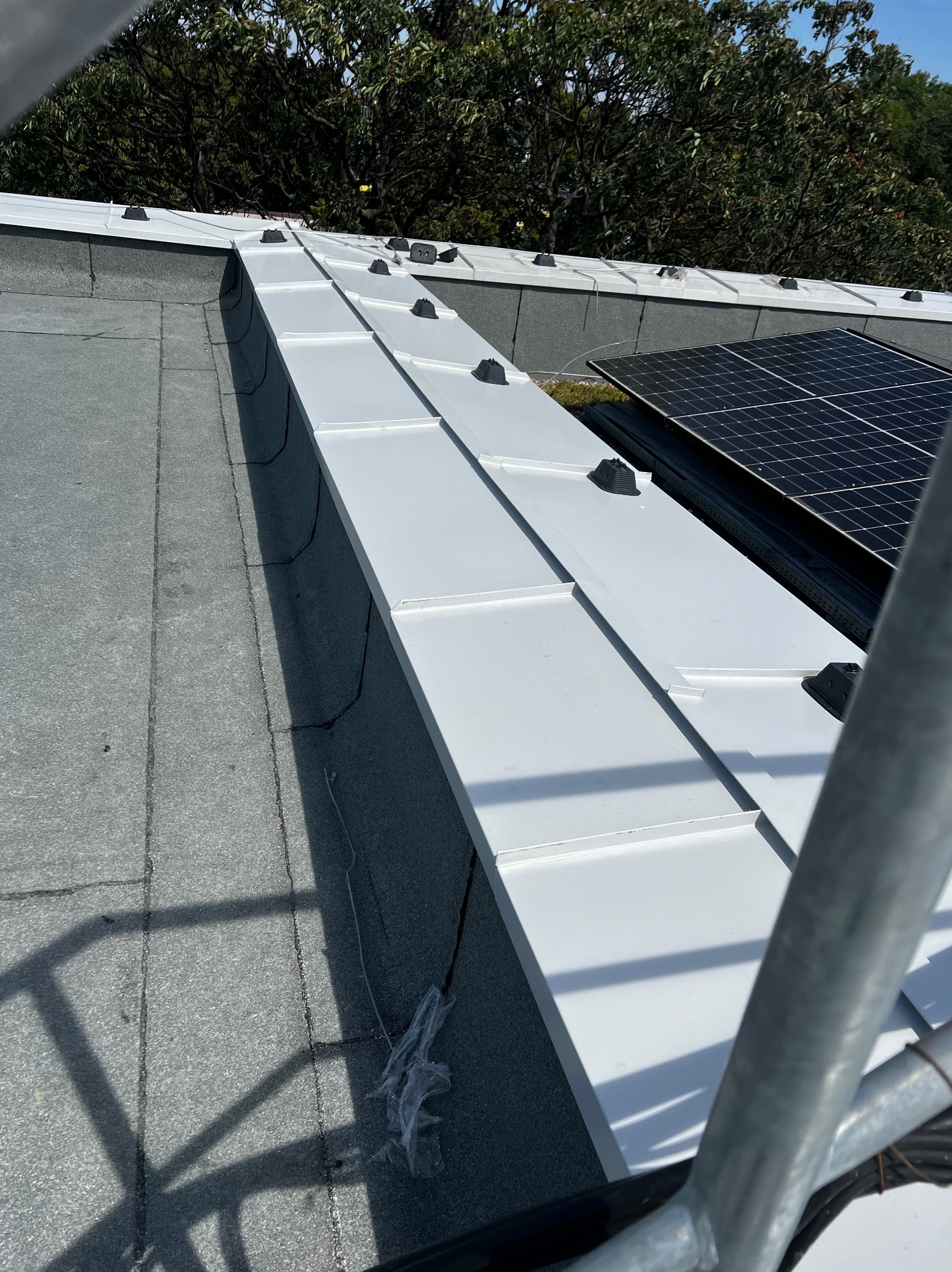 Nowoczesny dach z białymi panelami blachy na rąbek stojący, czarne elementy mocujące, widoczne panele fotowoltaiczne i fragment rusztowania.