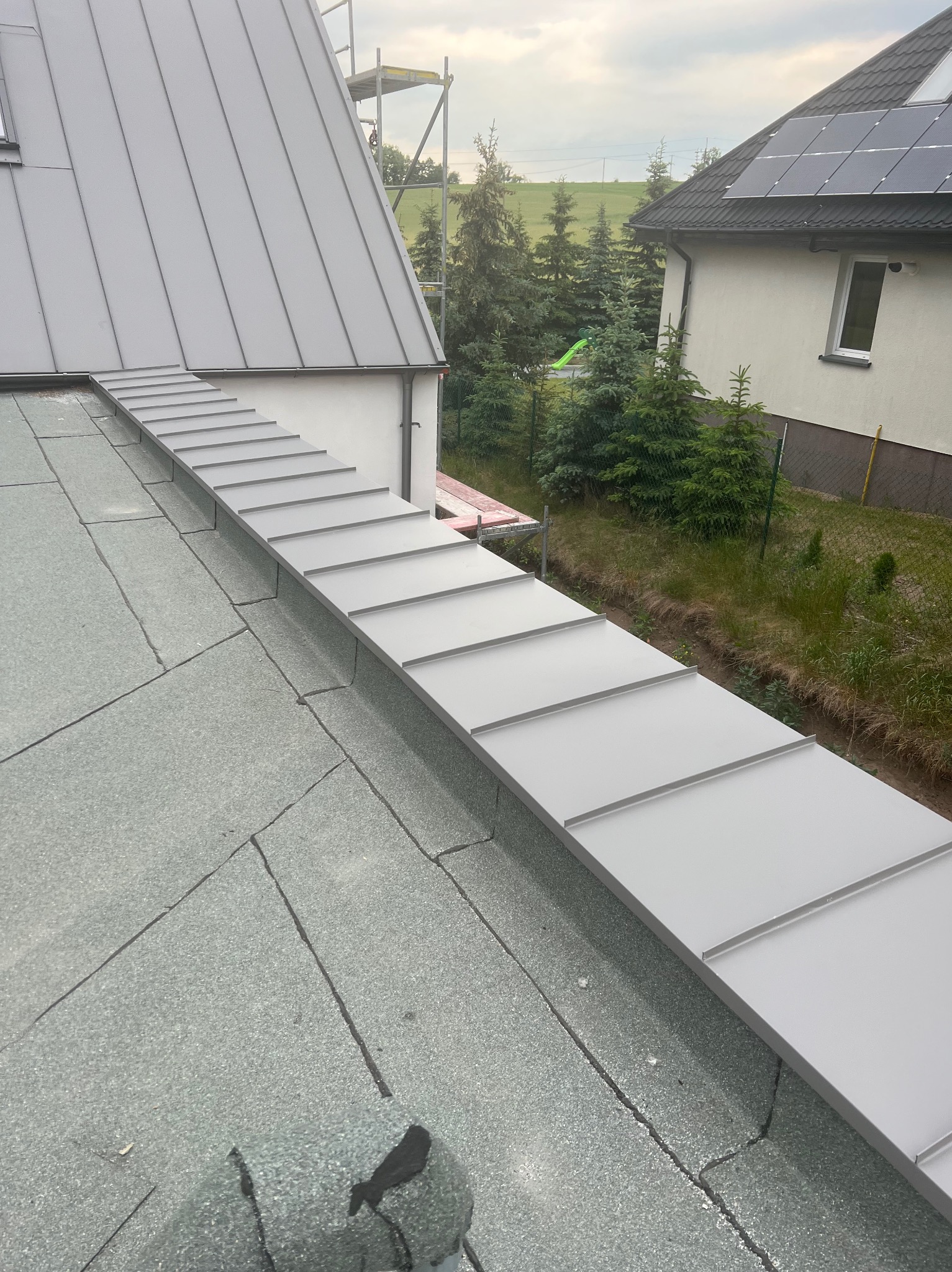 Szara obróbka blacharska na styku dachu pokrytego papą i ściany budynku, widok z góry na tle zieleni i sąsiedniego domu z panelami słonecznymi.