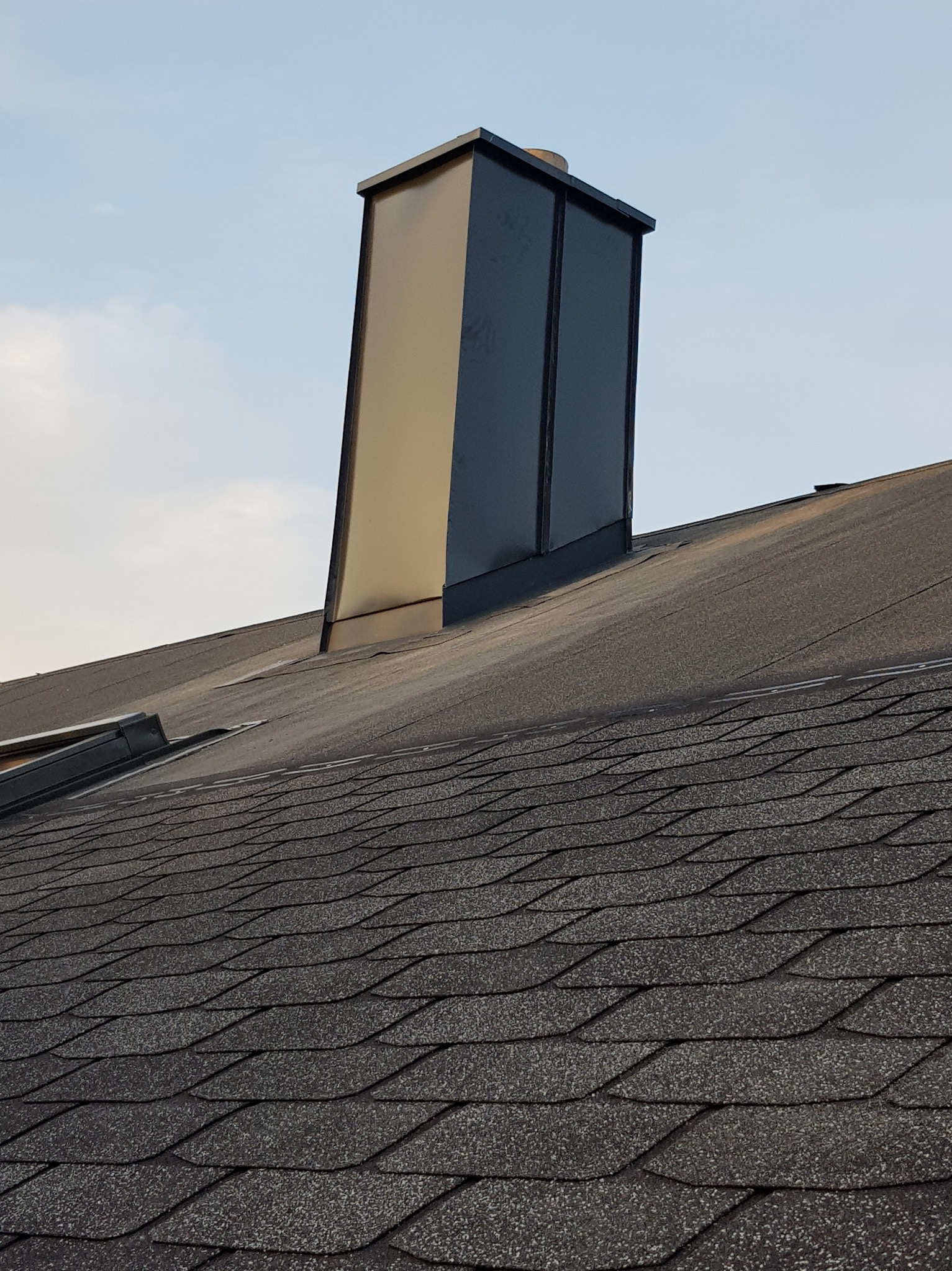 Nowy, błyszczący komin z obróbką blacharską na dachu pokrytym gontem bitumicznym w odcieniach szarości, widoczne łączenie z połacią dachu.