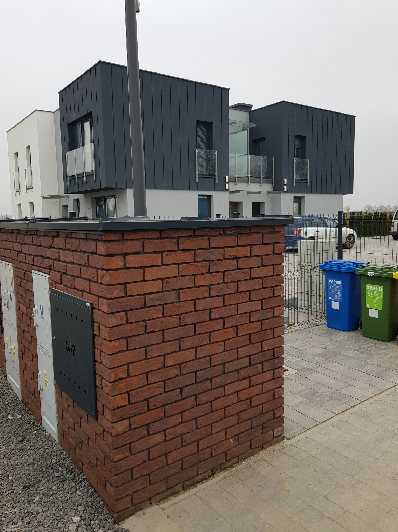 Nowoczesny dom z grafitowym dachem z blachy na rąbek stojący, widok z poziomu chodnika z kostki brukowej, na pierwszym planie mur z czerwonej cegły klinkierowej z szafkami licznikowymi i skrzynką...