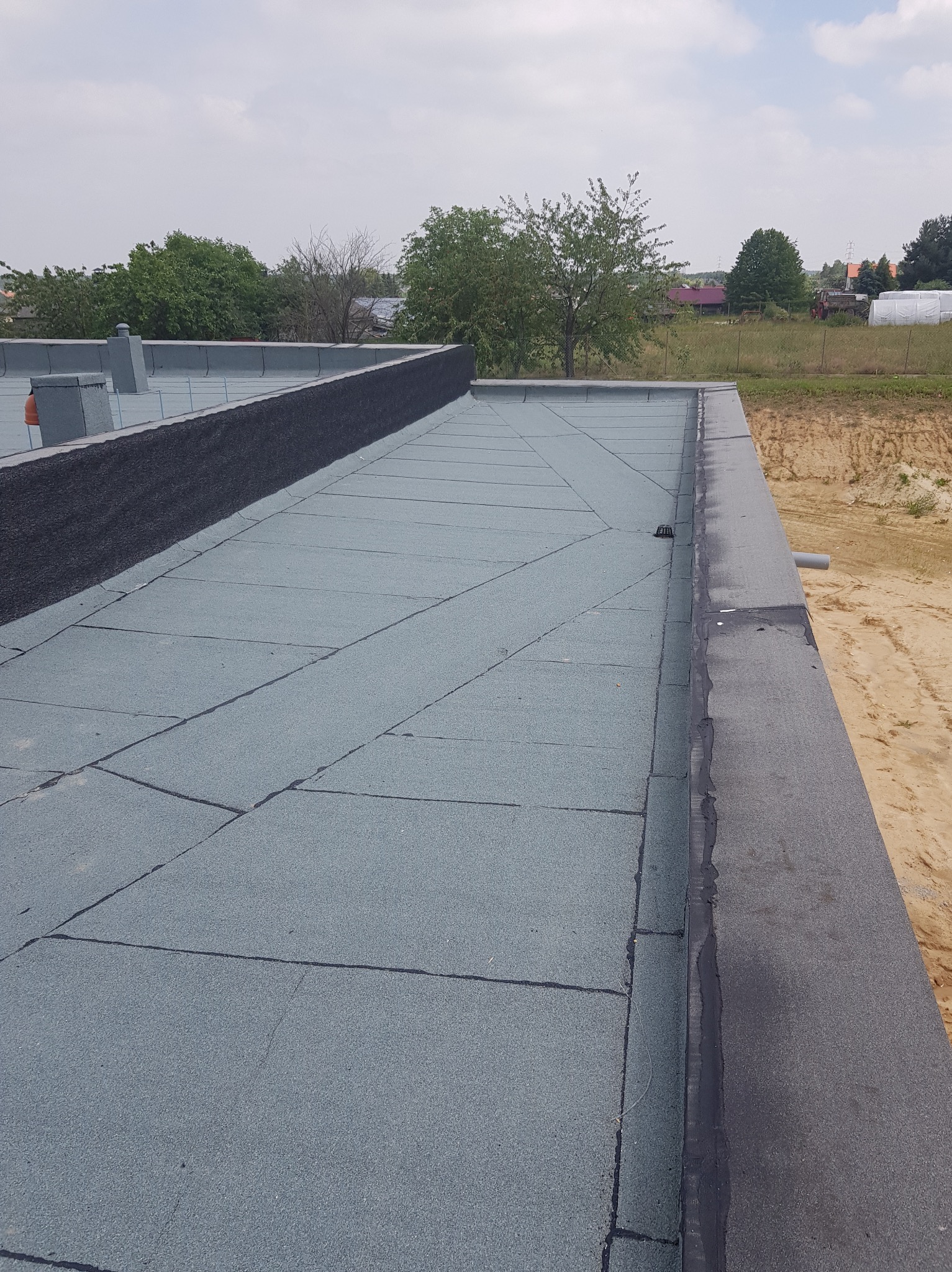 Płaski dach pokryty szarą papą bitumiczną w prostokątne płyty, widoczne kominy wentylacyjne i obróbka krawędzi dachu, w tle drzewa i zabudowania.