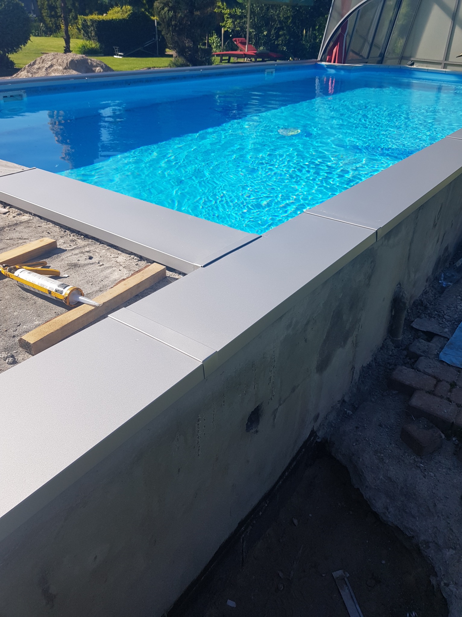 Montaż szarego obrzeża basenu z widocznym silikonem, drewnianymi rozpórkami i czystą, błękitną wodą w tle.