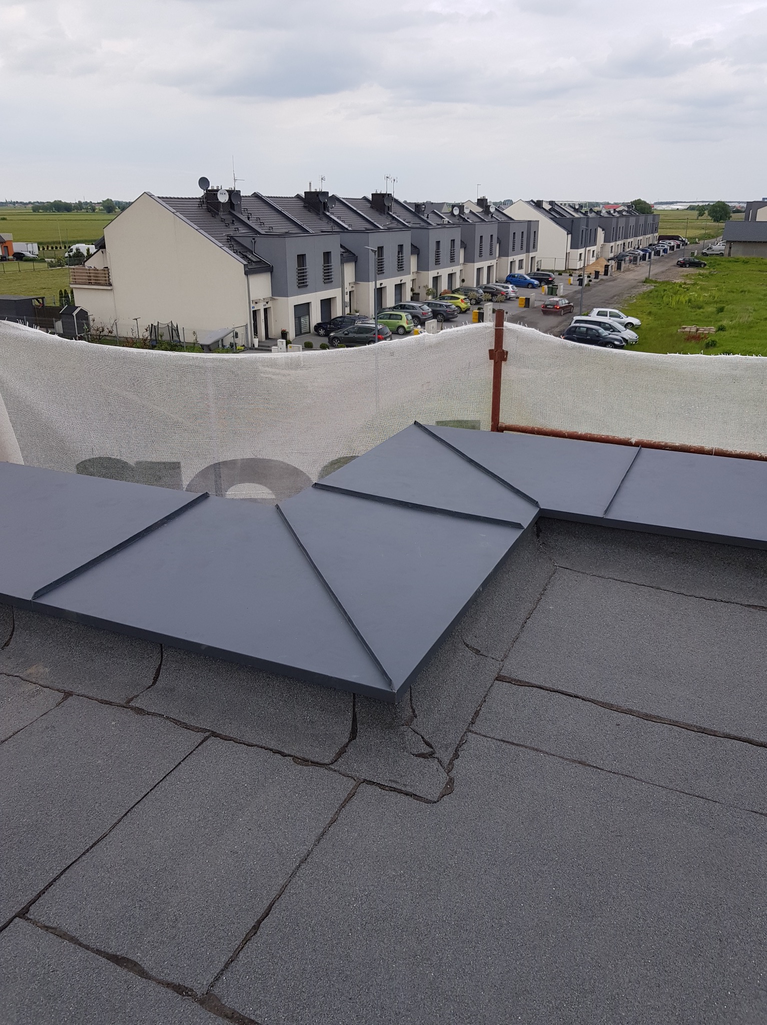 Szary dach z papy z obróbkami blacharskimi w kształcie rombów na tle szeregu domów jednorodzinnych i pochmurnego nieba.