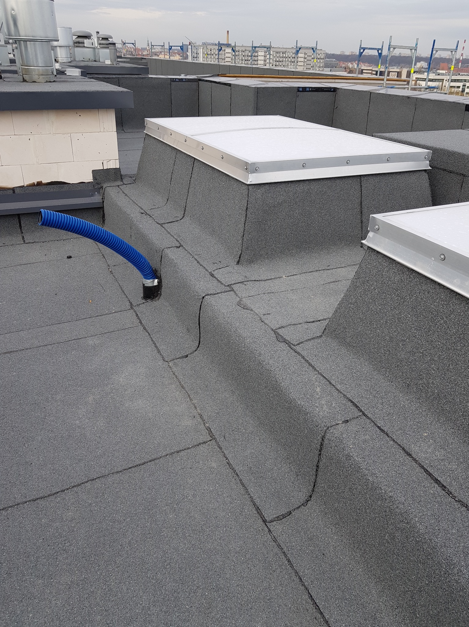 Wykończony dach pokryty papą termozgrzewalną z świetlikami dachowymi i niebieskim przewodem wentylacyjnym w Poznaniu, widok z góry.