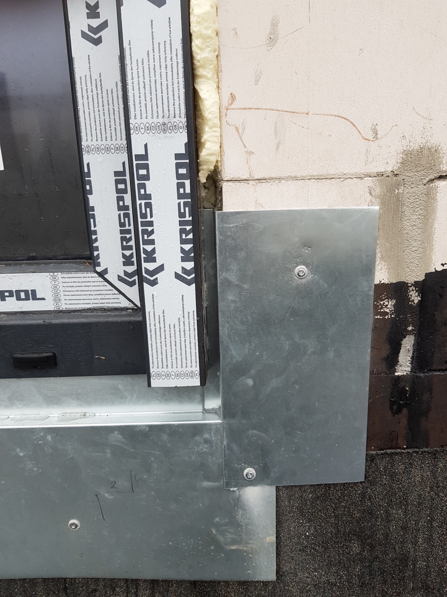 Szczegółowe ujęcie obróbki blacharskiej okna na dachu z widocznymi śrubami mocującymi i pianką uszczelniającą, fragment dachu pokrytego papą oraz ramą okna z folią ochronną producenta Krispol.