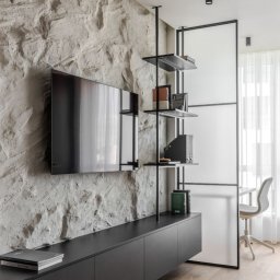 Patryk Gawrych Pracownia Dekoru - Ściana z dekoracyjną fakturą kamienia, na niej zawieszony telewizor. Poniżej czarna szafka RTV, obok regał z metalowymi półkami i przeszklona ścianka działowa oddzielająca przestrzeń biurową...