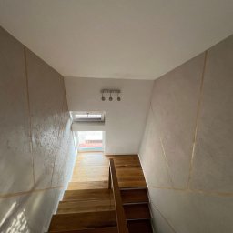 Patryk Gawrych Pracownia Dekoru - Klatka schodowa z drewnianymi schodami i ścianami wykończonymi dekoracyjną fakturą z delikatnymi złotymi liniami, oświetlona naturalnym światłem wpadającym przez okno.