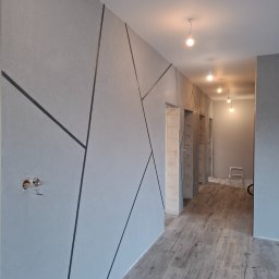 Patryk Gawrych Pracownia Dekoru - Korytarz w szarościach z dekoracyjnymi, geometrycznymi liniami na ścianie, widoczne niedokończone gniazdko elektryczne i drewnopodobna podłoga.