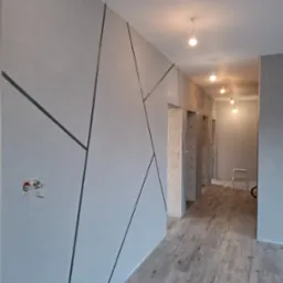 Korytarz w szarościach z dekoracyjnymi, geometrycznymi liniami na ścianie, widoczne niedokończone gniazdko elektryczne i drewnopodobna podłoga.