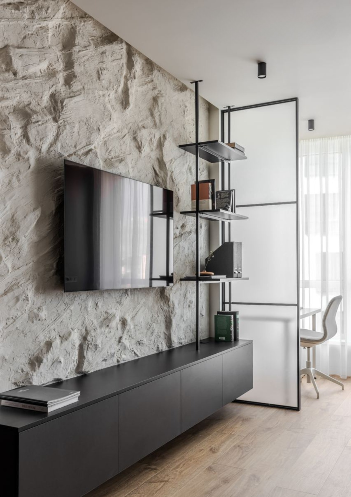 Ściana z dekoracyjną fakturą kamienia, na niej zawieszony telewizor. Poniżej czarna szafka RTV, obok regał z metalowymi półkami i przeszklona ścianka działowa oddzielająca przestrzeń biurową...