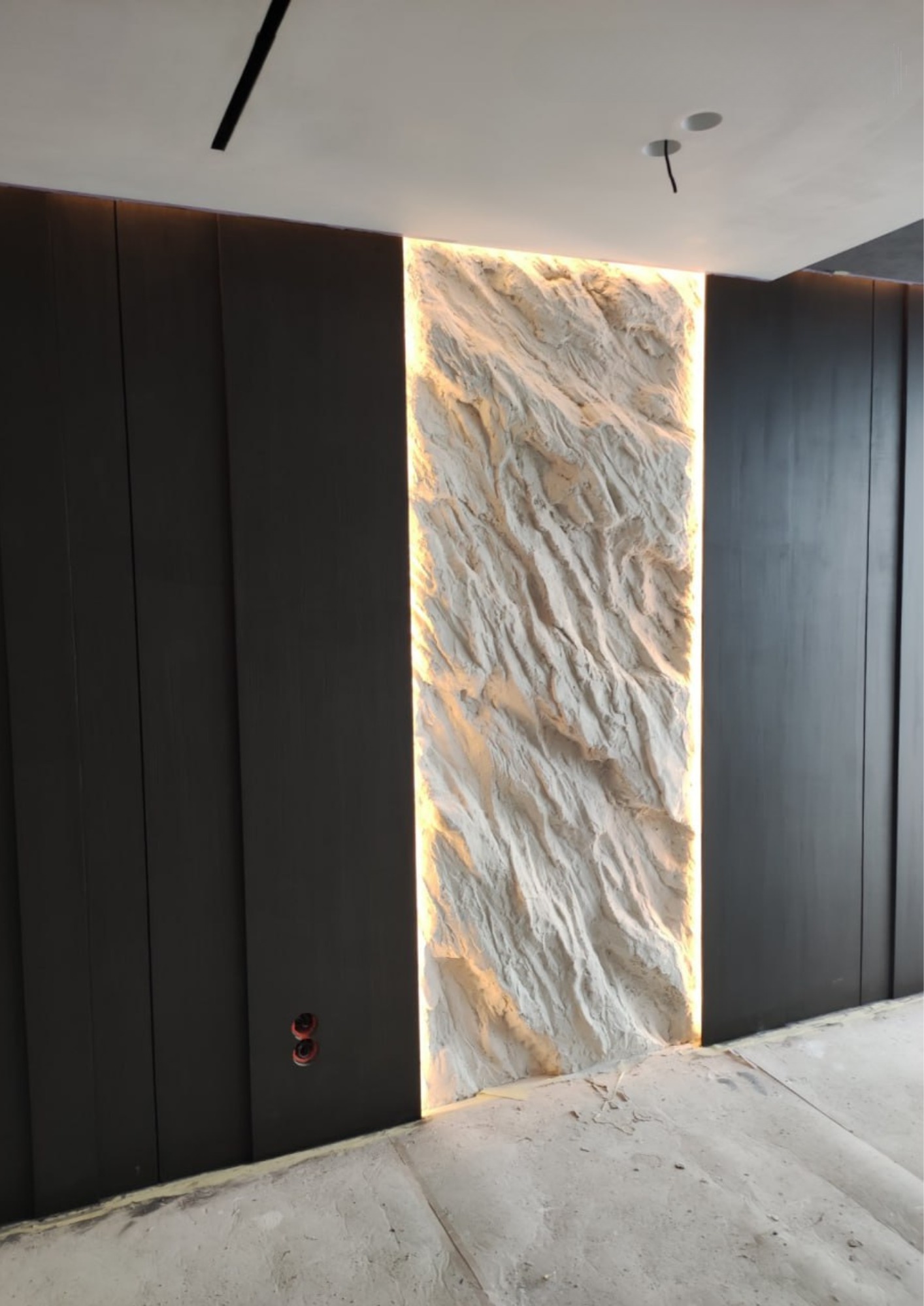 Dekoracyjna ściana z efektem trójwymiarowej skały, podświetlona od tyłu ciepłym światłem, sąsiadująca z ciemnymi, pionowymi panelami ściennymi.