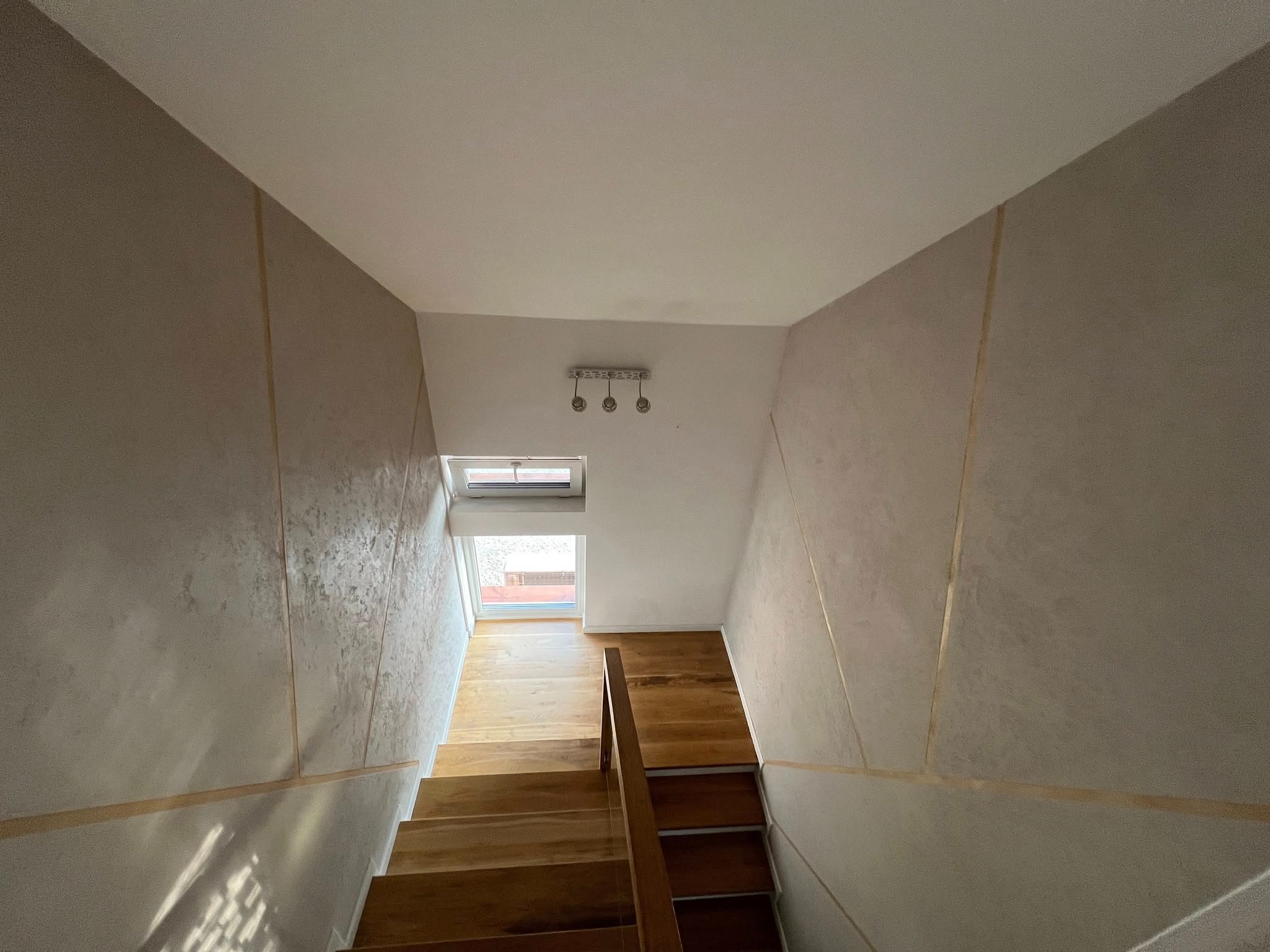 Klatka schodowa z drewnianymi schodami i ścianami wykończonymi dekoracyjną fakturą z delikatnymi złotymi liniami, oświetlona naturalnym światłem wpadającym przez okno.