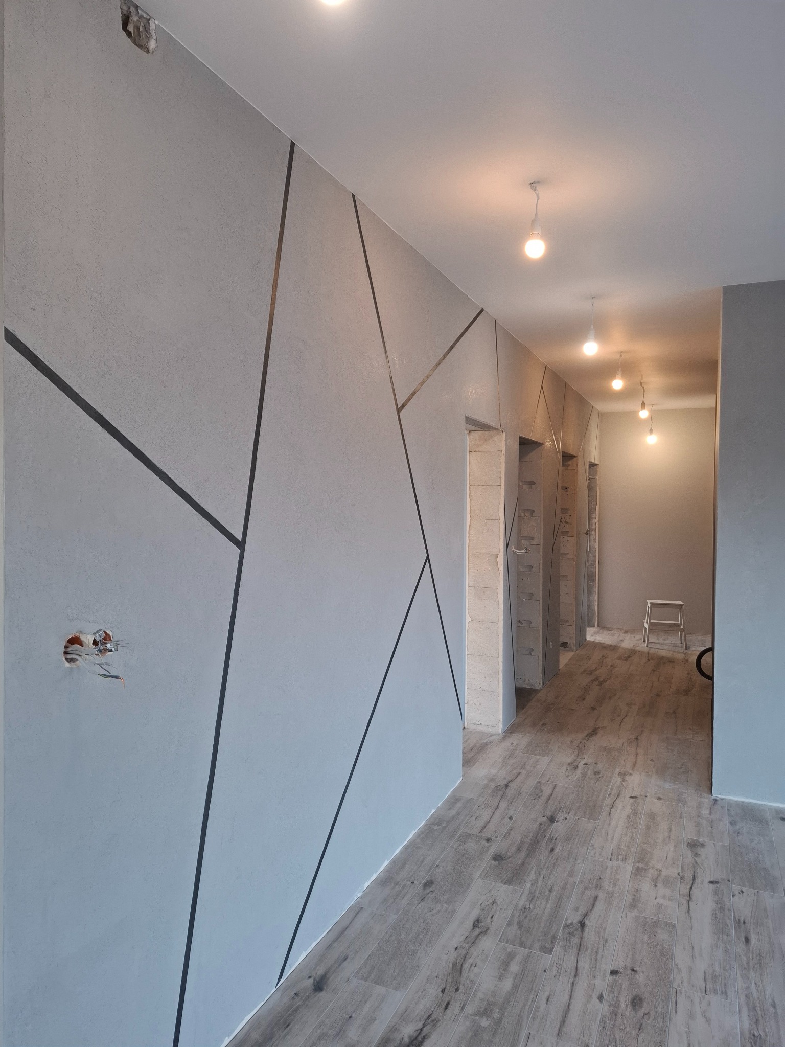 Korytarz w szarościach z dekoracyjnymi, geometrycznymi liniami na ścianie, widoczne niedokończone gniazdko elektryczne i drewnopodobna podłoga.