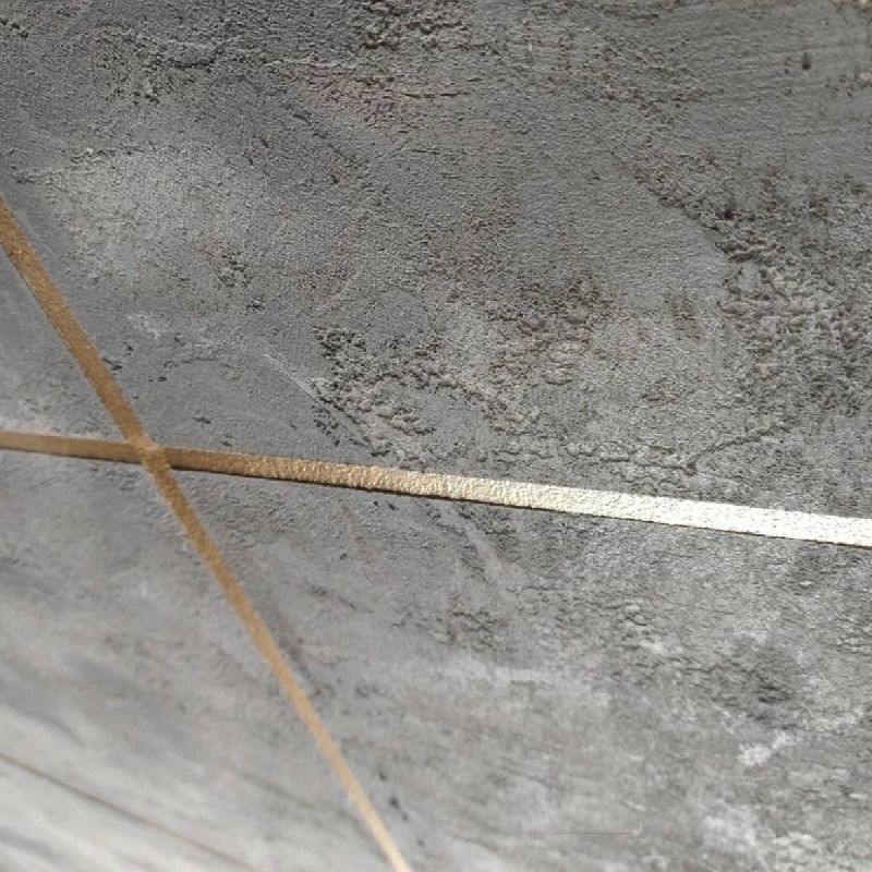 Szara, teksturowana ściana z dekoracyjnymi, złotymi paskami przecinającymi się pod kątem prostym, tworzącymi geometryczny wzór.