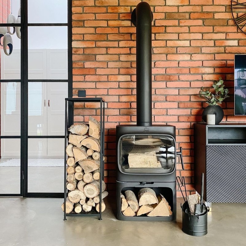 Nowoczesny piec kominkowy z czarnym kominem na tle ściany z czerwonej cegły, obok stojak na drewno i akcesoria kominkowe.