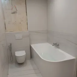 Biała, nowoczesna łazienka w trakcie remontu: widoczna wanna z wodą, podwieszana toaleta, płytki imitujące marmur oraz fragment ściany w stanie surowym z wystającymi kablami.