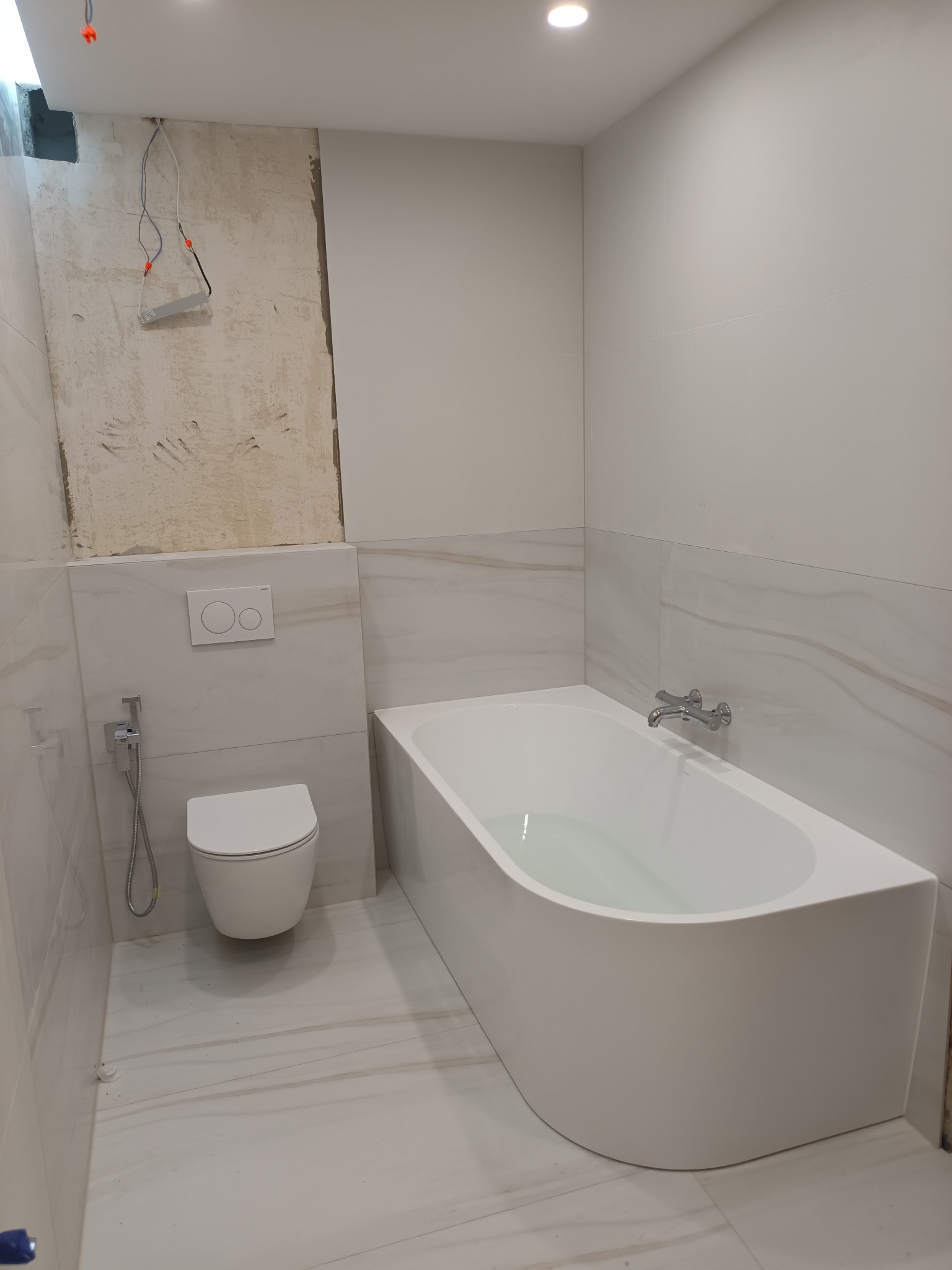 Biała, nowoczesna łazienka w trakcie remontu: widoczna wanna z wodą, podwieszana toaleta, płytki imitujące marmur oraz fragment ściany w stanie surowym z wystającymi kablami.
