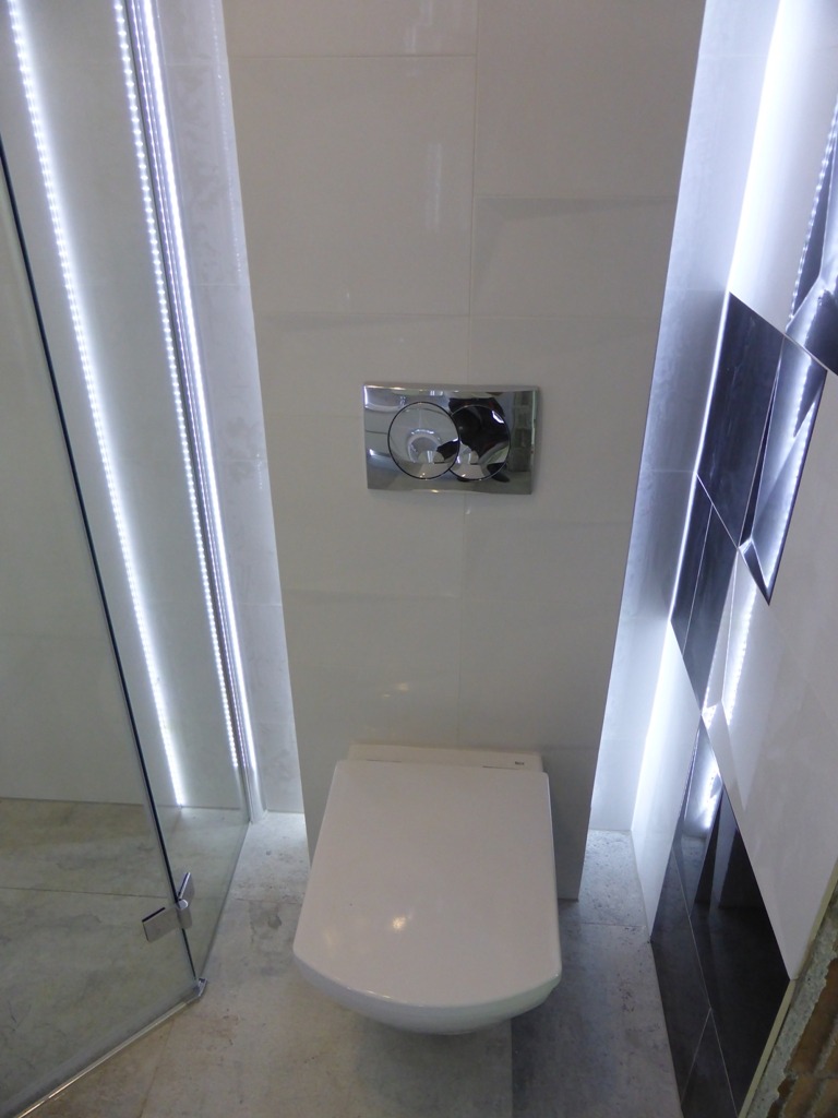 Nowoczesna toaleta podwieszana z chromowanym przyciskiem spłukującym, oświetleniem LED wzdłuż ścian i kabiny prysznicowej, oraz dekoracyjnymi czarno-białymi płytkami po prawej stronie.