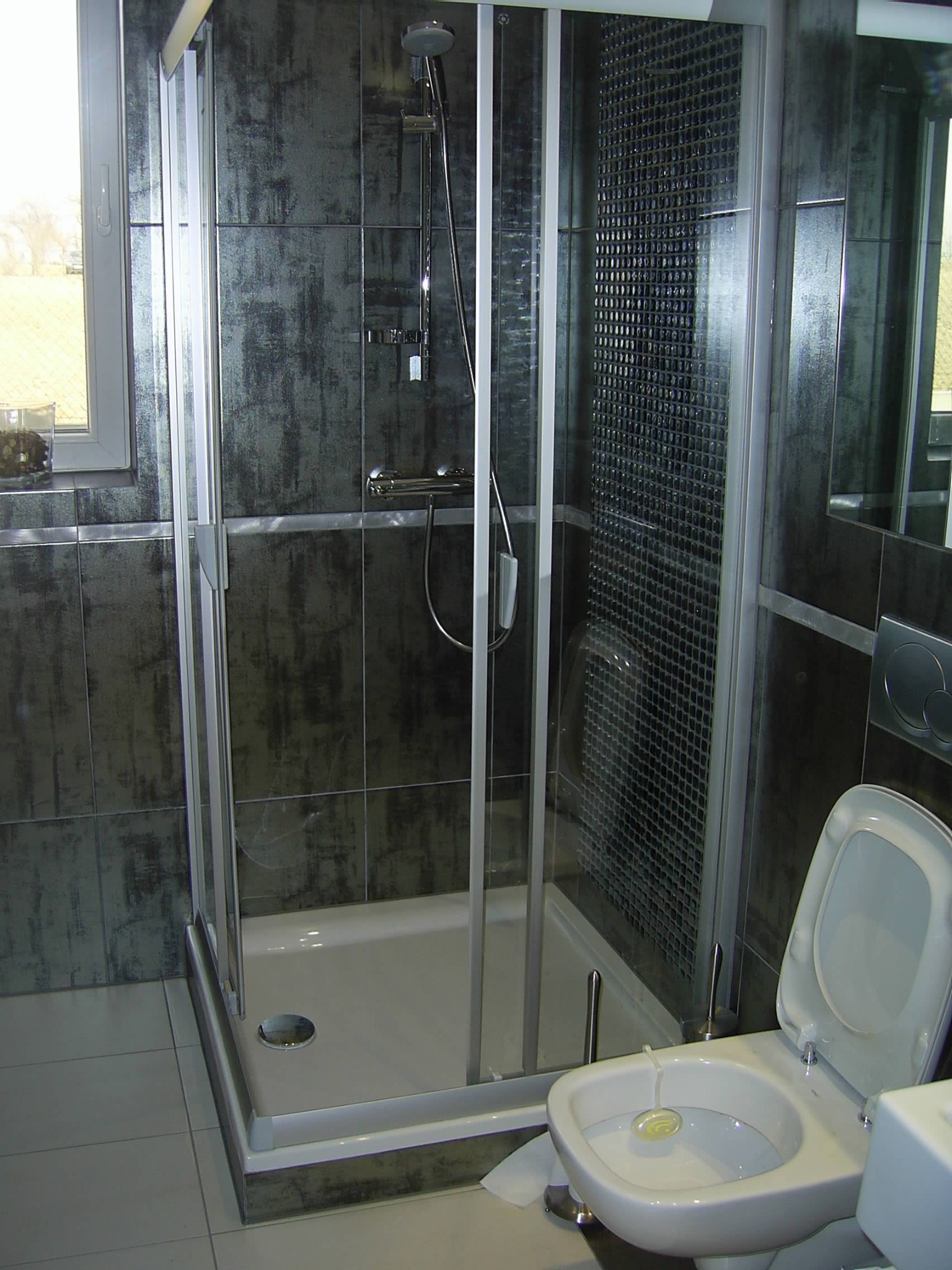 Kabina prysznicowa z przesuwnymi drzwiami i szarymi płytkami imitującymi beton, widoczny również bidet z odświeżaczem w misce, w tle okno.