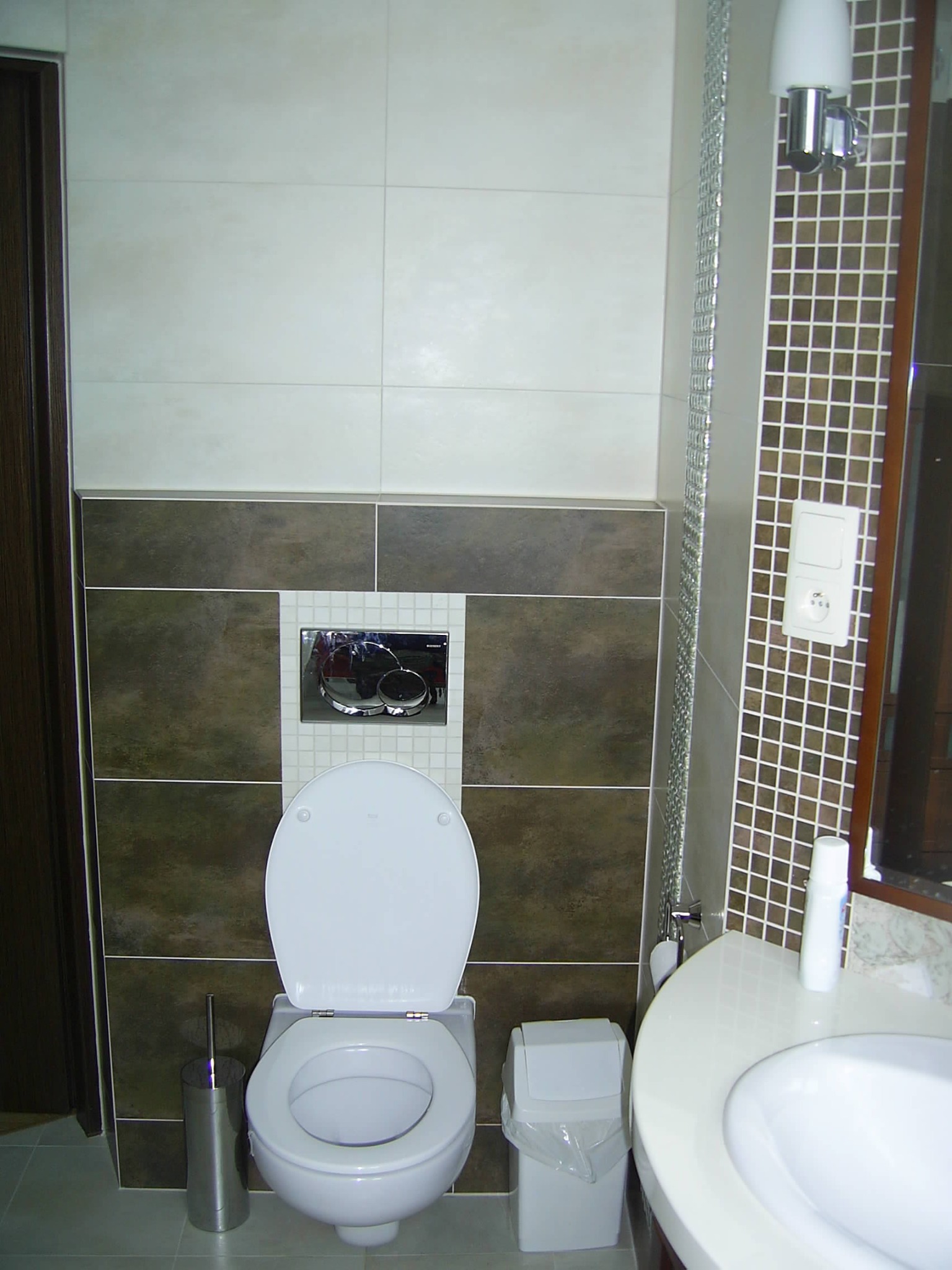 Wykończona łazienka z brązowo-białymi płytkami, białą ceramiką i chromowanymi detalami, widok na sedes z otwartą klapą, umywalkę oraz kosz na śmieci z widoczną folią.