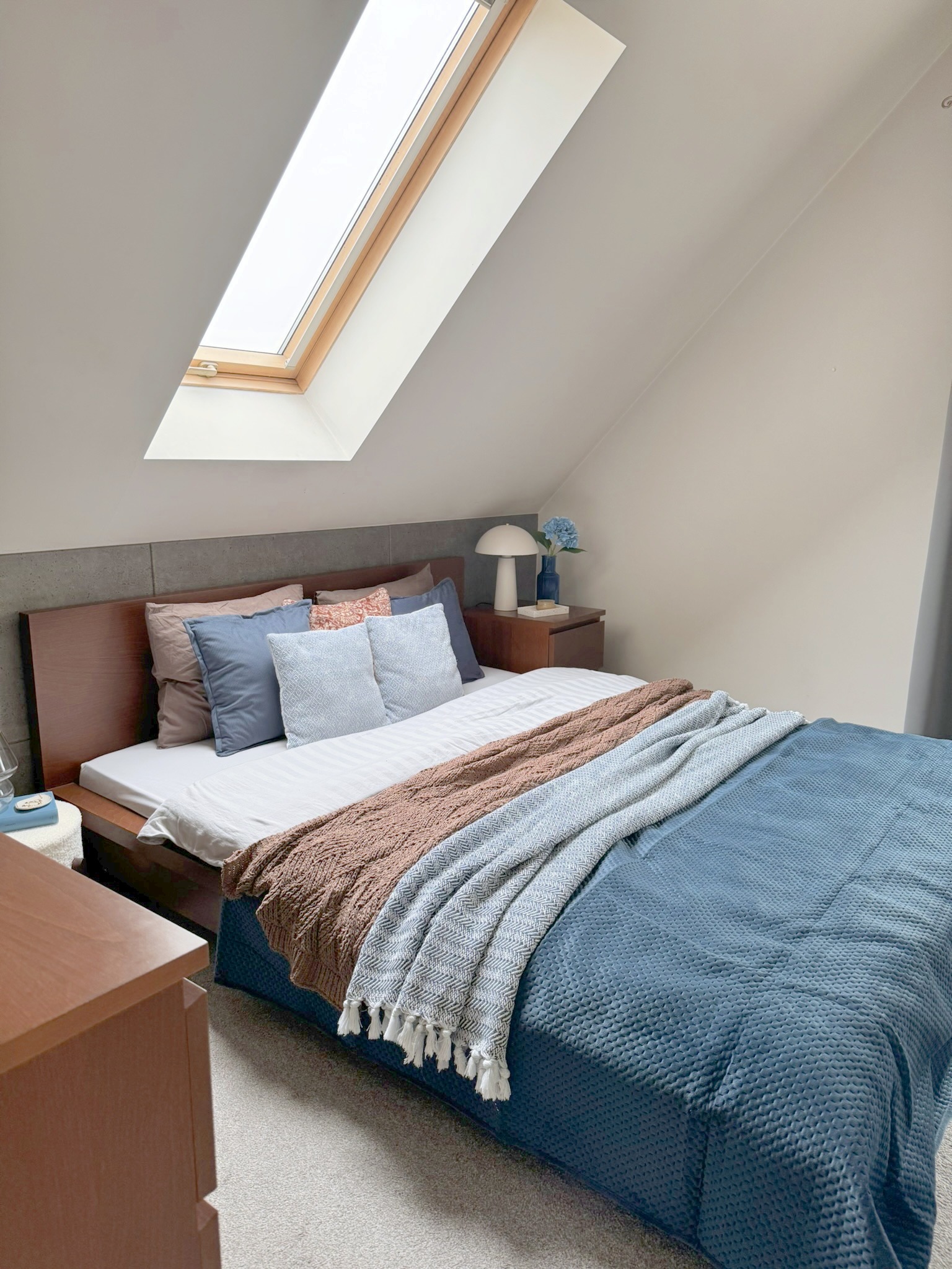 Przytulna sypialnia na poddaszu z łóżkiem, pledami w warkocze i oknem dachowym wpuszczającym światło. Stonowana kolorystyka i minimalistyczny wystrój.
