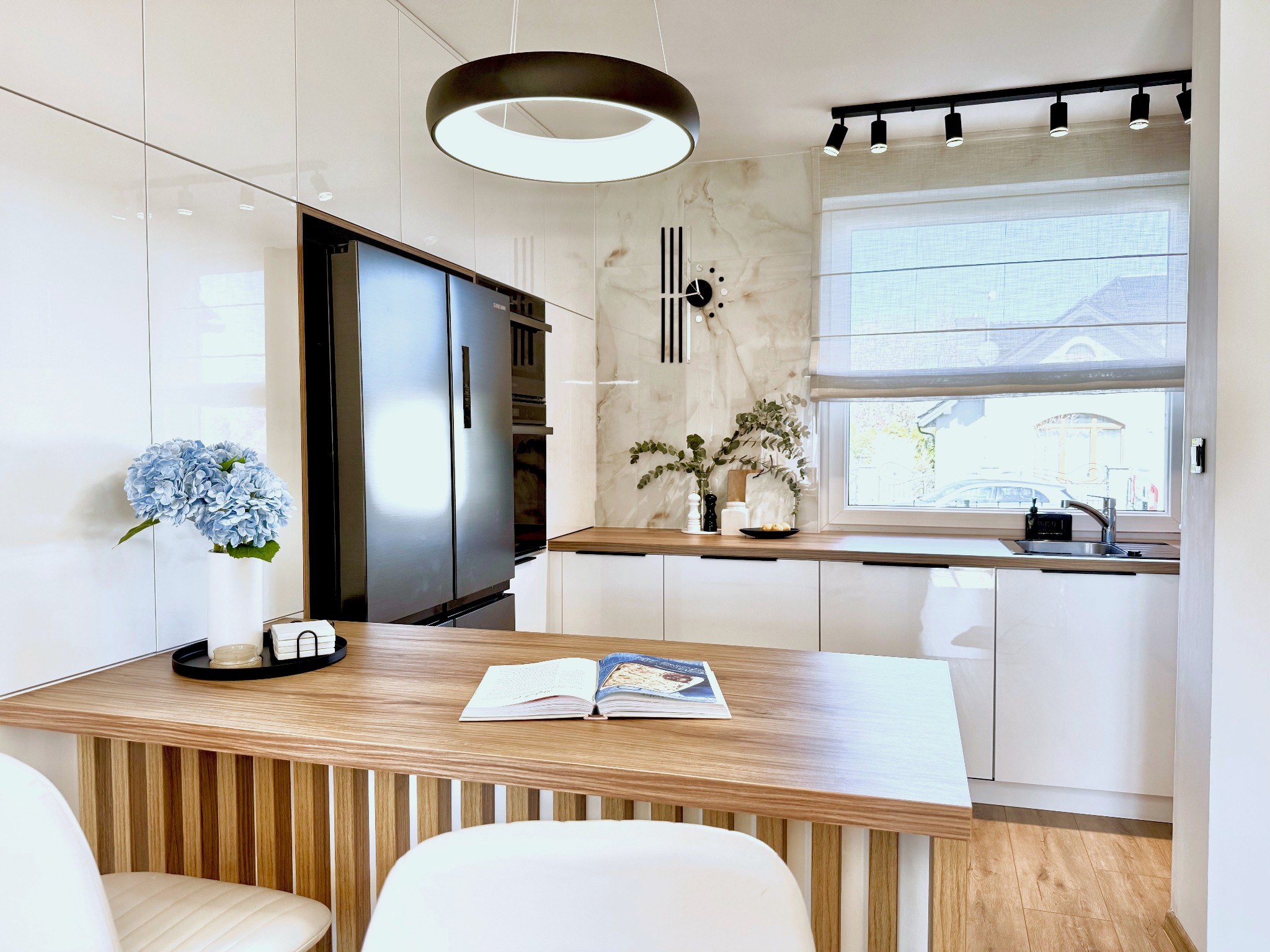 Jasna kuchnia z białymi szafkami i drewnianym blatem, okrągła lampa sufitowa, kwiaty w wazonie, otwarta książka kucharska na wyspie. Nowoczesny design.
