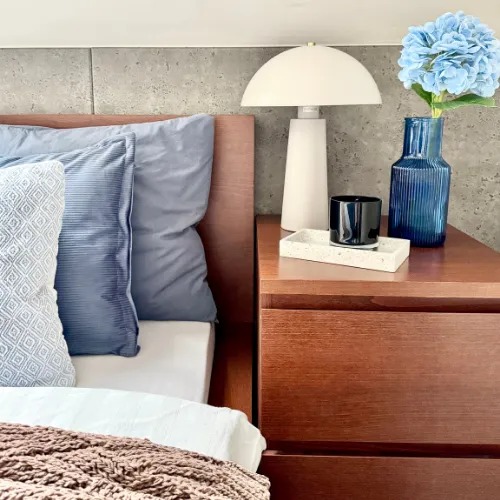 Sypialnia z drewnianym stolikiem nocnym, lampą i niebieskim wazonem z kwiatami. Przytulny kącik nocny z detalami dekoracyjnymi.