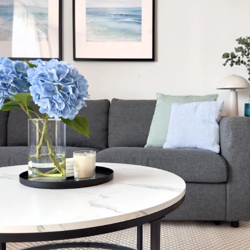 Jasne wnętrze: szary narożnik z poduszkami, okrągły stolik z marmurowym blatem, wazon z niebieskimi hortensjami i świeca na tacy.