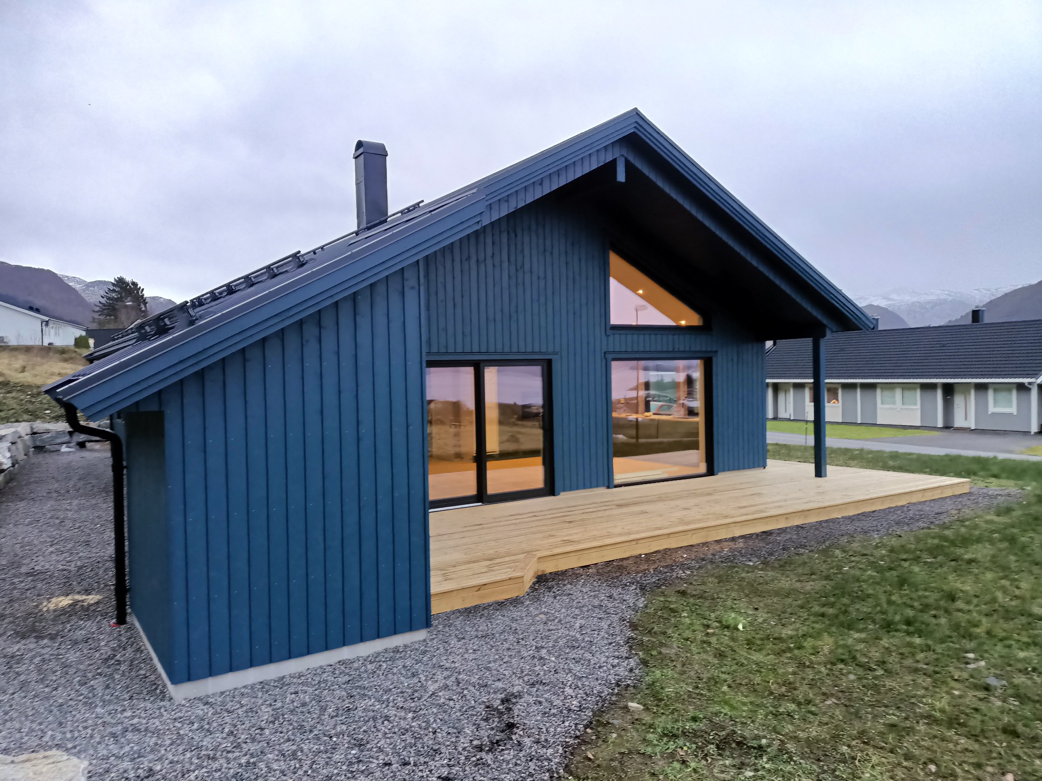 Niebieski dom szkieletowy z drewnianym tarasem i dużymi oknami, widok z zewnątrz w pochmurny dzień, na tle gór.