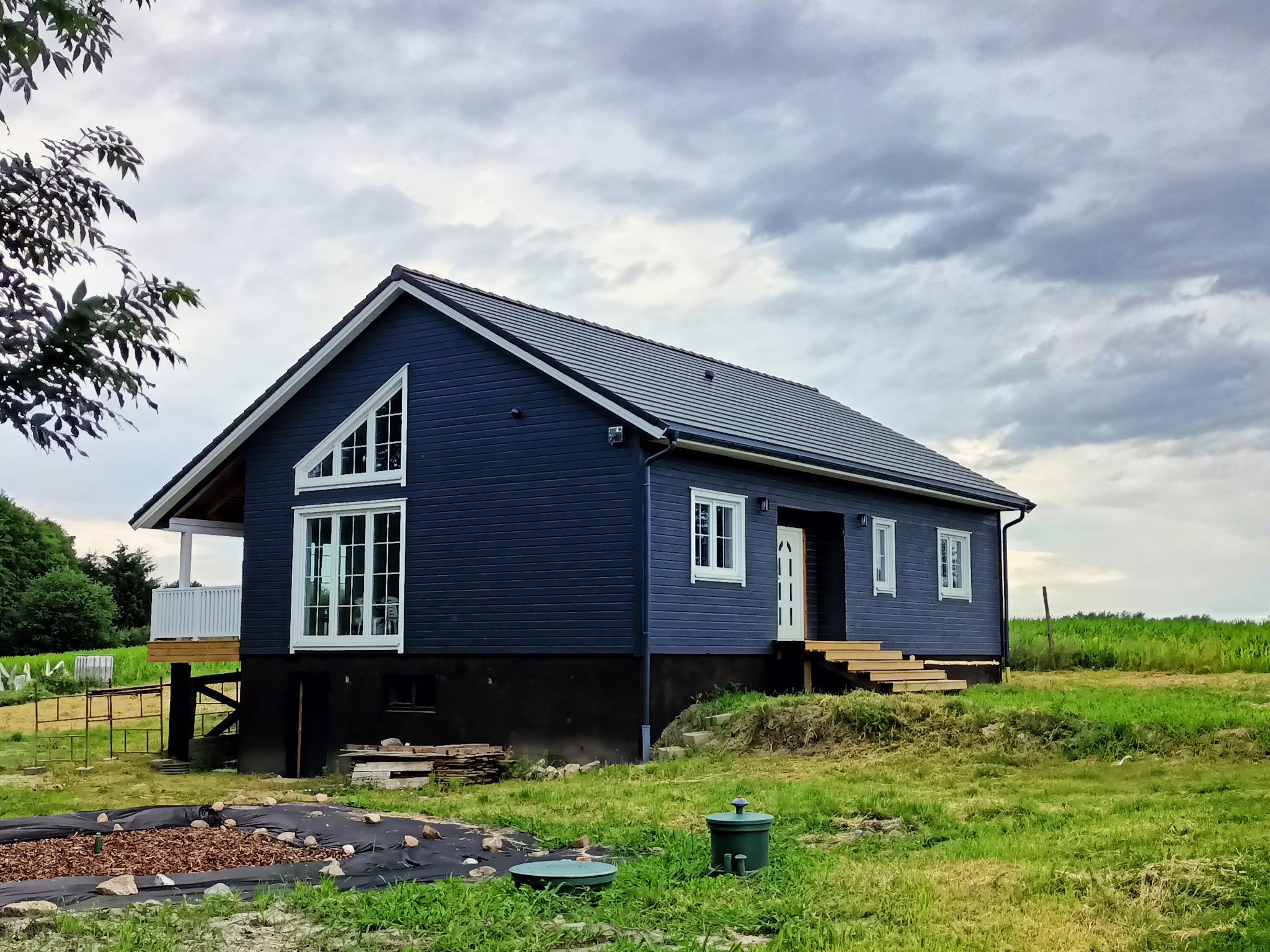 Niebieski dom szkieletowy z białymi oknami i schodami wejściowymi, częściowo na podwyższeniu, otoczony zielonym trawnikiem i pochmurnym niebem.