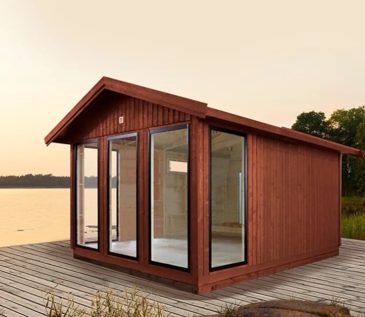 Niewykończona, drewniana konstrukcja z dużymi oknami na drewnianym pomoście nad wodą, potencjalnie altana lub domek letniskowy w trakcie budowy.