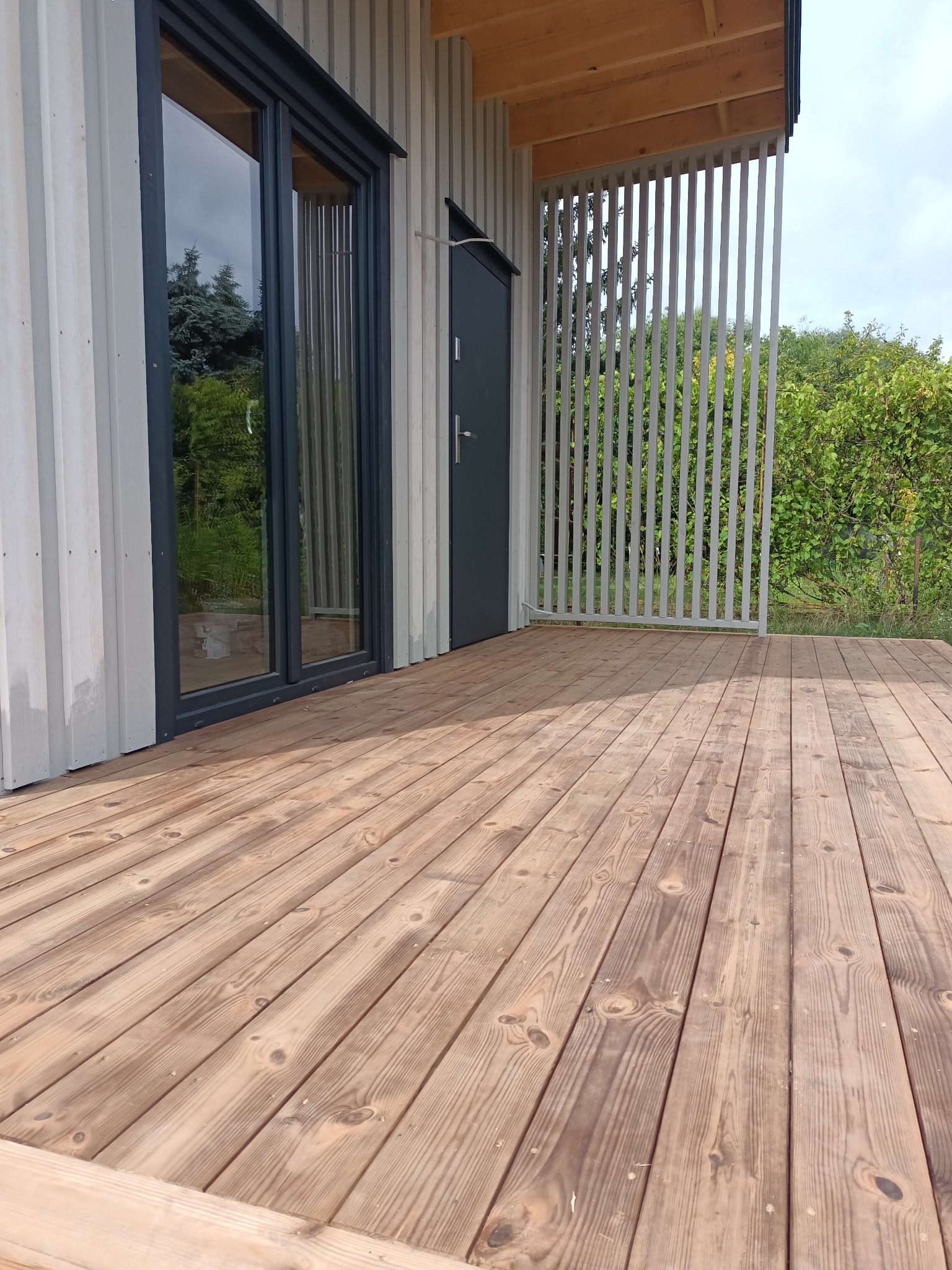 Nowoczesny taras z desek w odcieniach brązu, przylegający do budynku z szarymi elementami. Widoczne pionowe, białe lamele i zielona roślinność w tle.