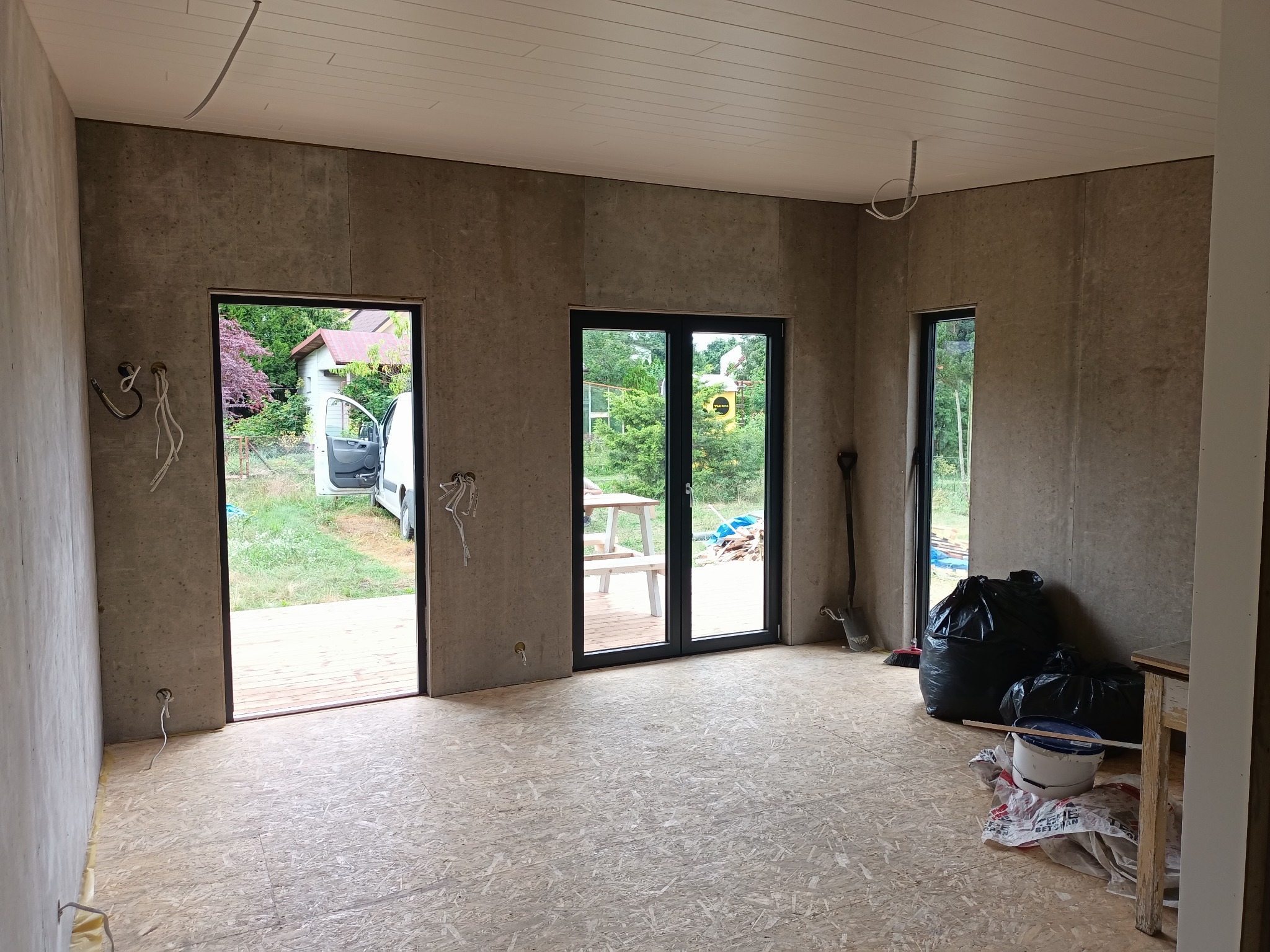 Wnętrze domu w trakcie budowy: surowe ściany, podłoga z płyt OSB, widoczne instalacje elektryczne, okna z widokiem na zieleń. W tle narzędzia i worki na śmieci.